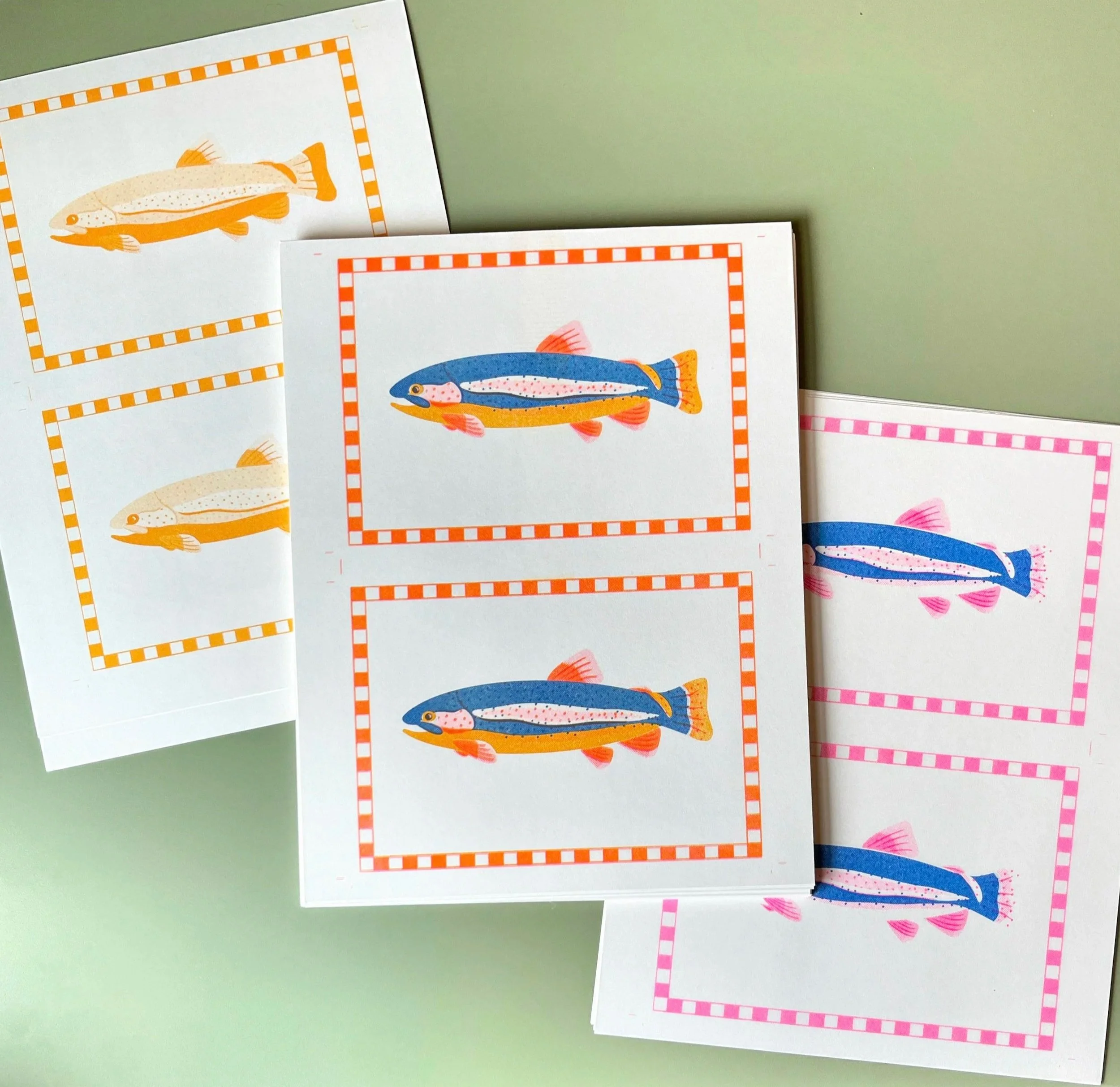 Flatlay of three risograph prints in different steps of the printing process. The prints are of two 5x7" rainbow trout illustrations printed on 8.5x11" paper.