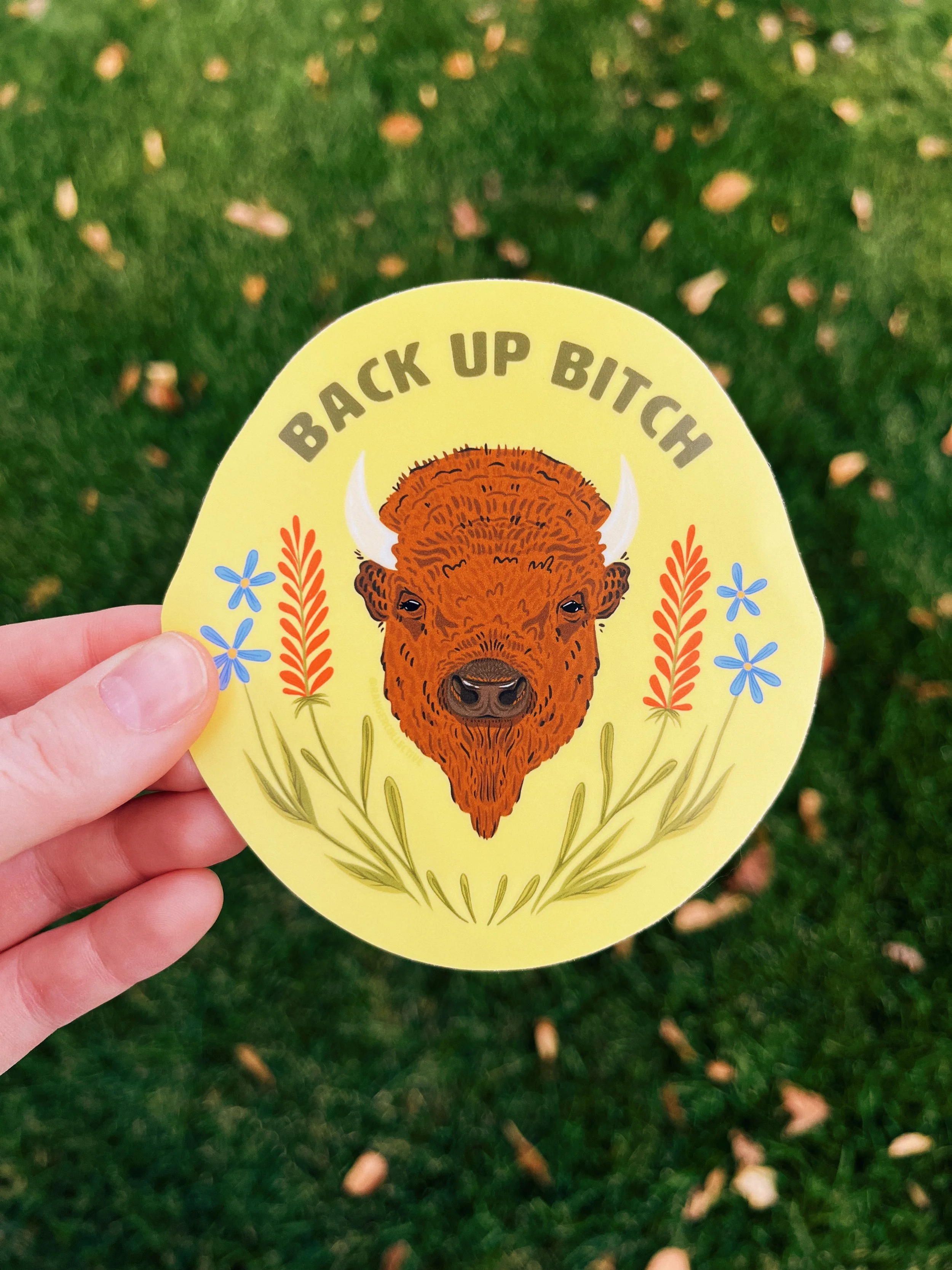 Image of a hand holding a sticker that has the face of a bison surrounded by wildflowers and reads "back up bitch" with a yellow background.