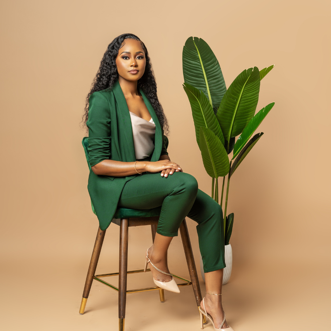A woman in a green suit and nude heels sitting on a wooden chair with a large green plant beside her, against a beige background.