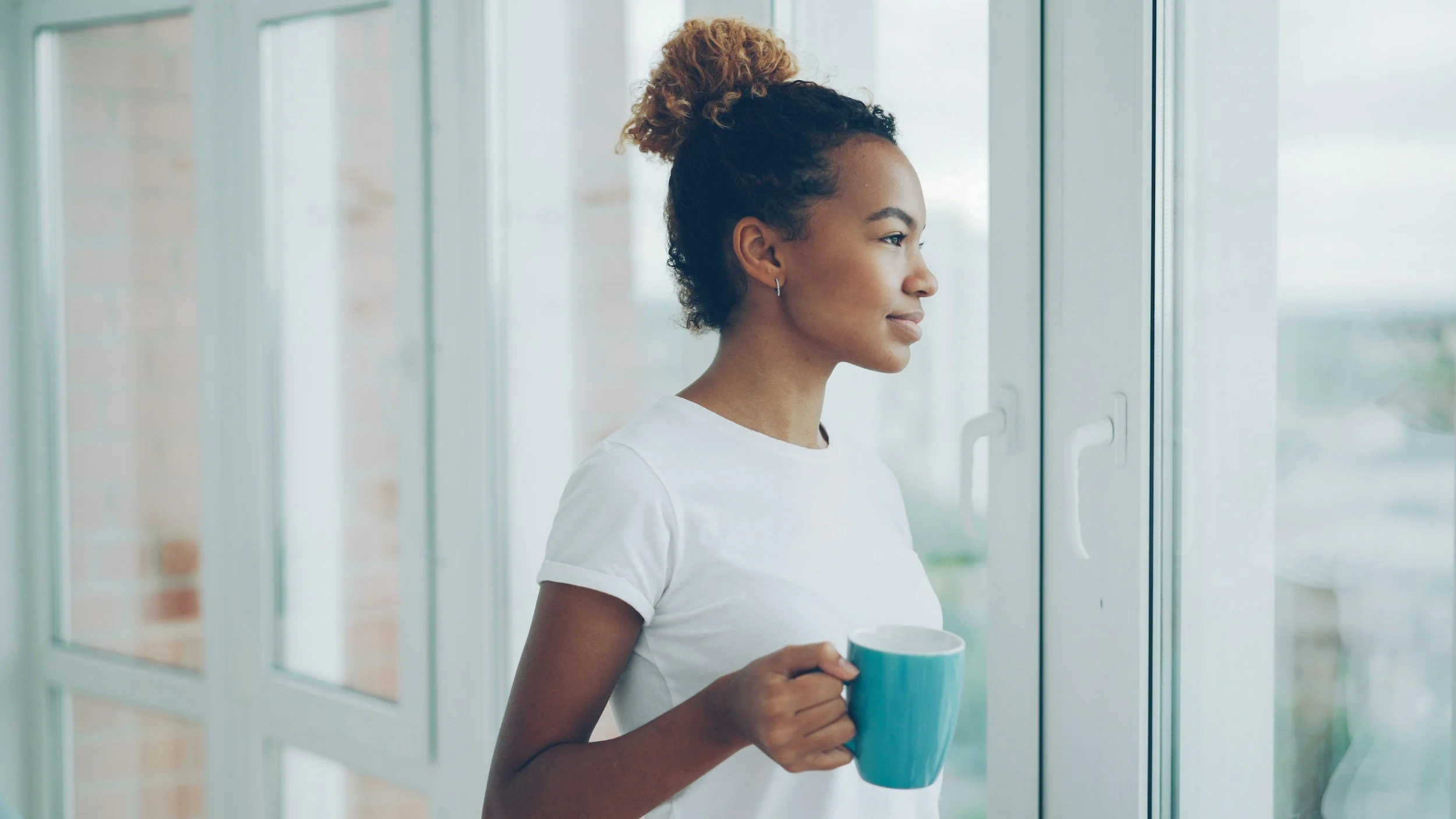 black woman looking out of window with blue mug in hand
