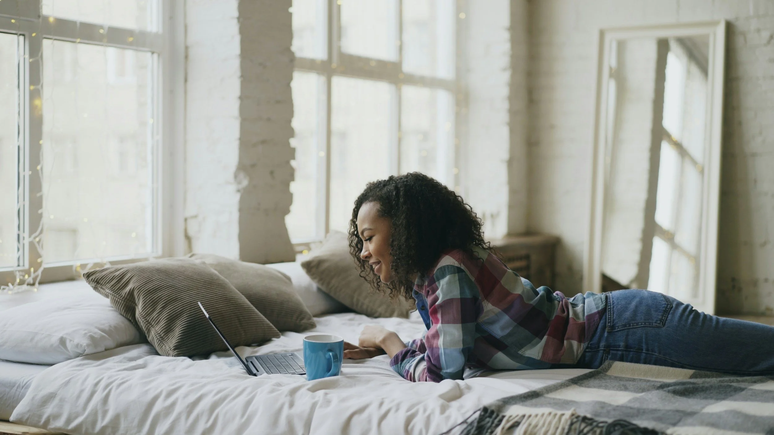 woman on bed looking at computer with coffee mug beside her on the bed