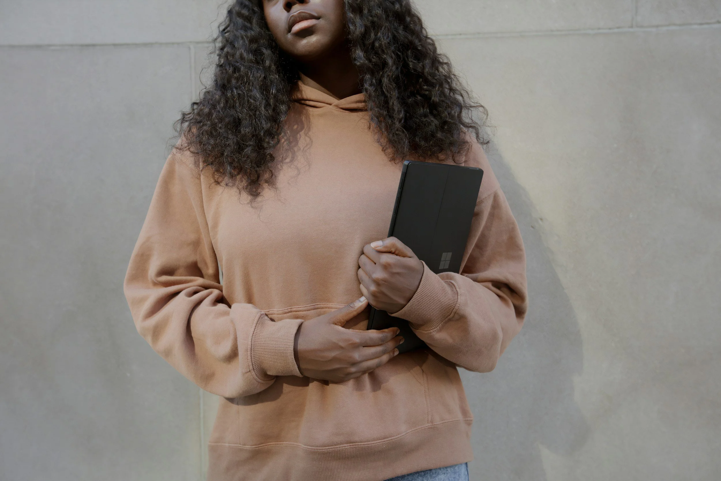 Woman standing with computer in hand with a brown hoodie on, looking away