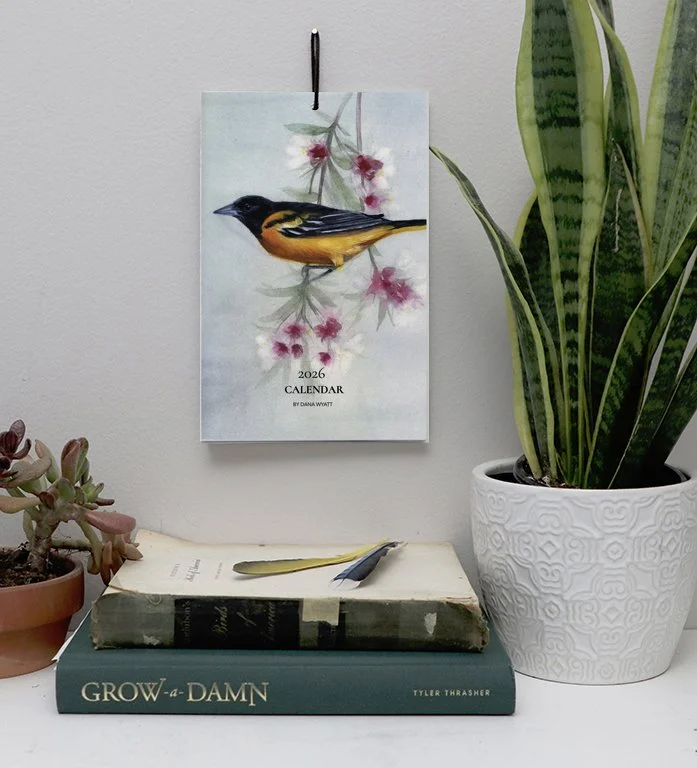 A 2026 calendar with an illustration of a bird perched on pink flowers, hanging on a wall, next to a large potted snake plant, with two books and a small succulent plant on a surface below.
