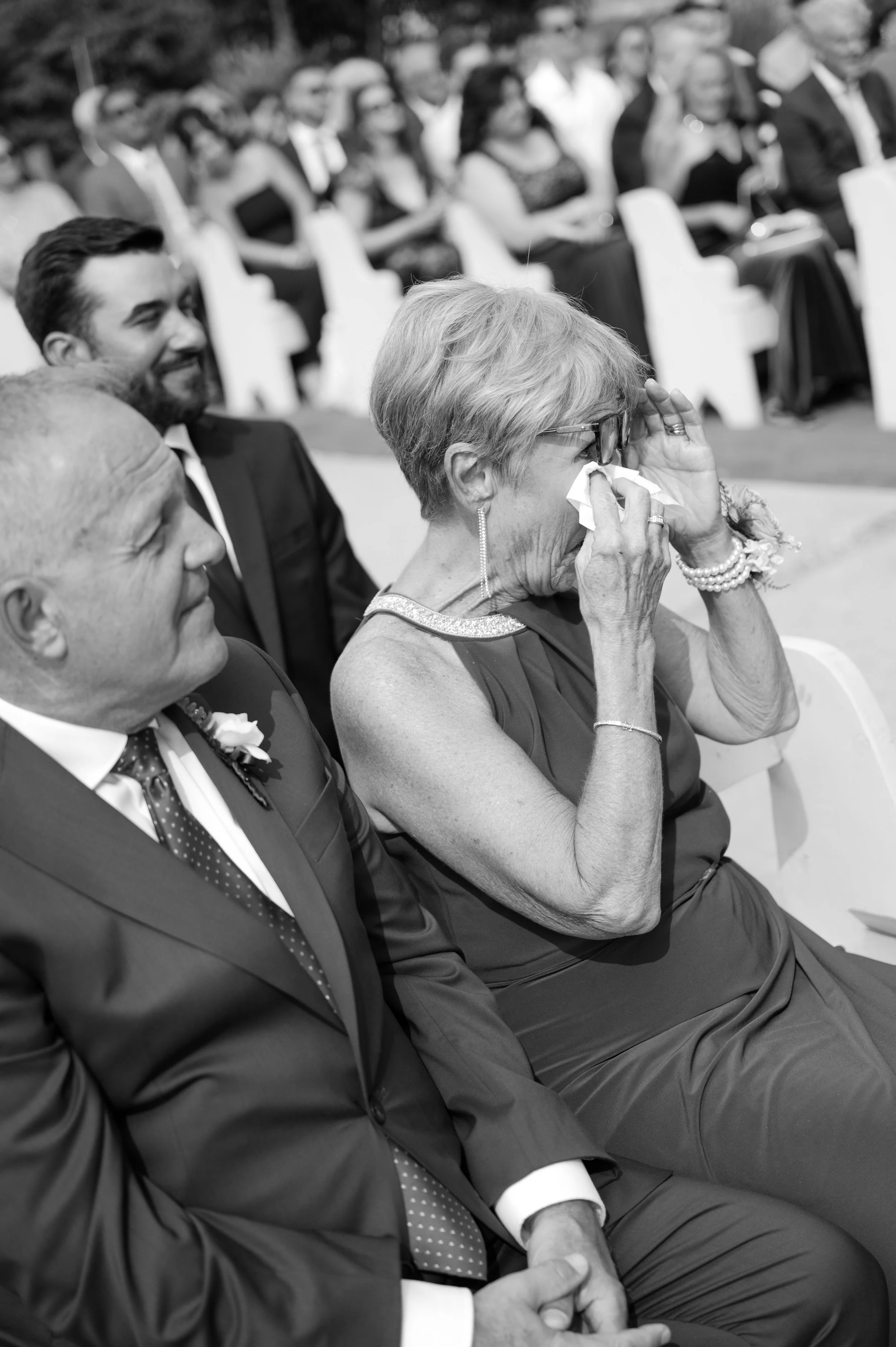 An elderly woman in a formal dress is wiping a tear away while seated at a formal event, surrounded by other well-dressed attendees.