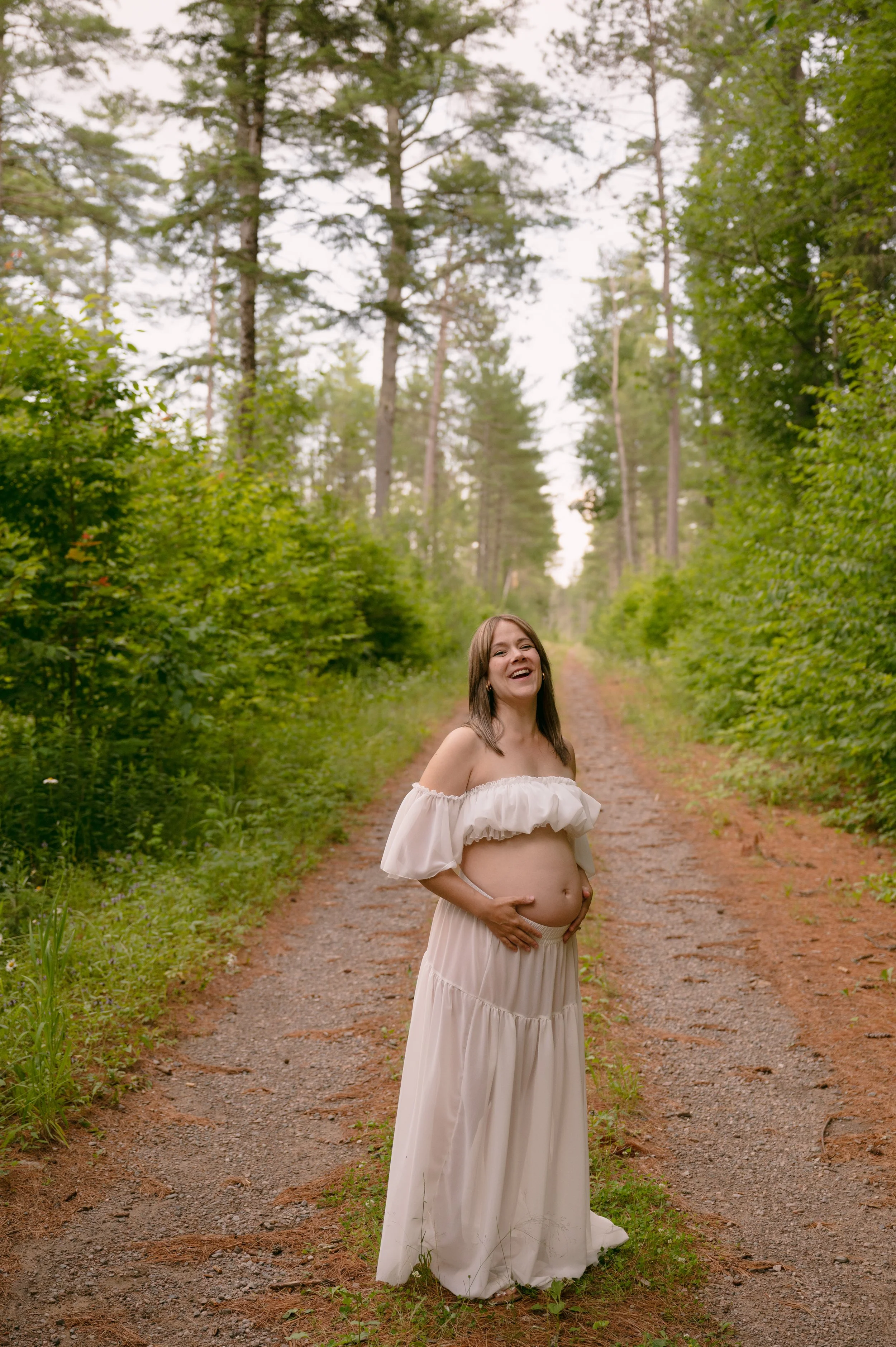 A pregnant woman smiling and holding her belly while standing on a dirt path in a forest.