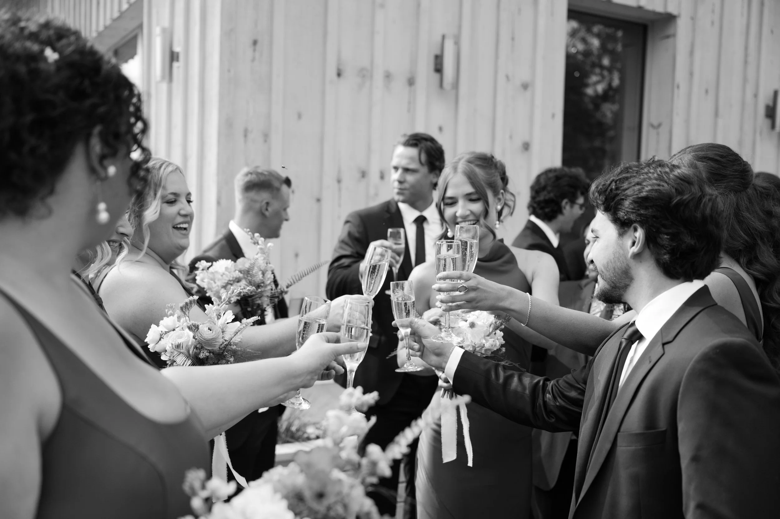 Group of people celebrating with champagne at a wedding reception, smiling and raising glasses in a toast outdoors with a modern wooden building in the background.