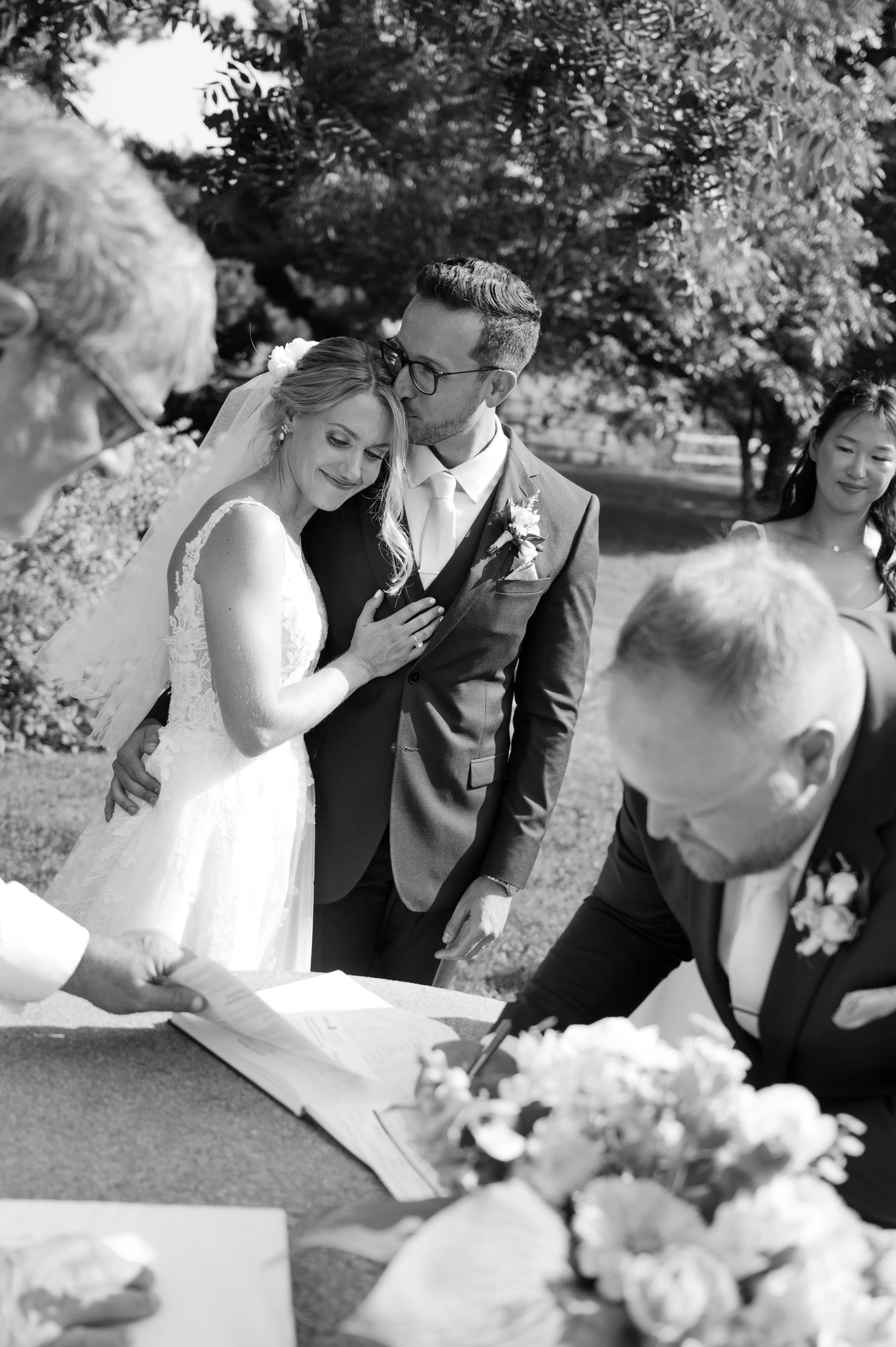 Black and white photo of a wedding ceremony outdoors with the bride and groom standing close, exchanging vows, surrounded by family and friends.