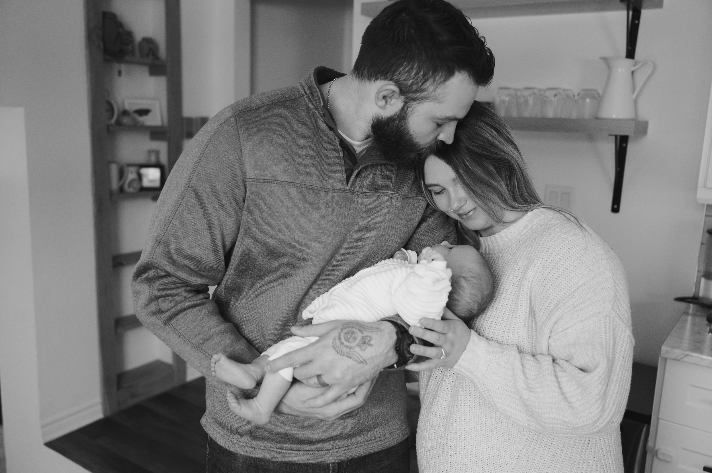 A couple holds their newborn baby in a hospital room, with the father kissing the mother's forehead.