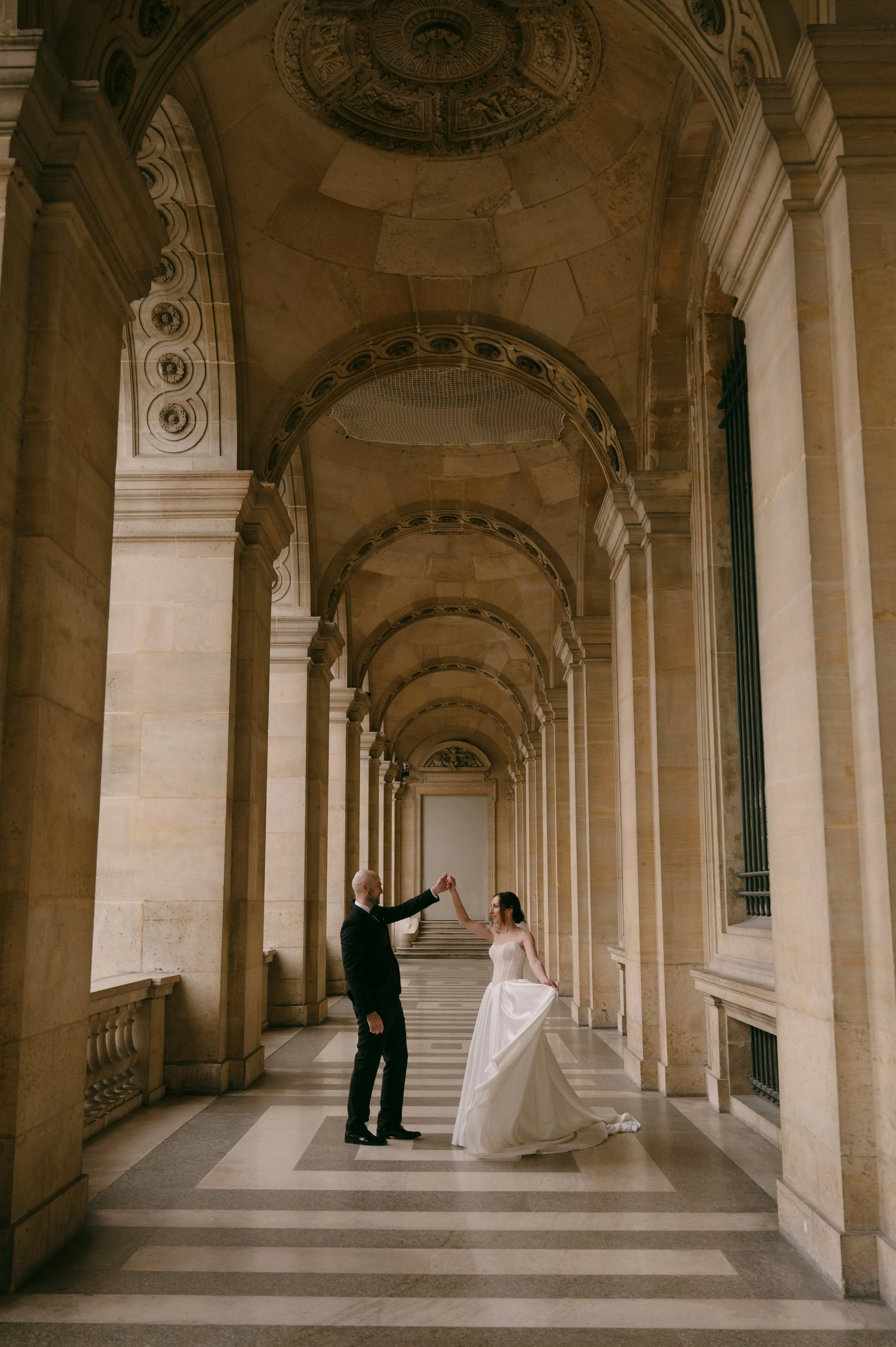 A bride and groom dancing in a grand, arched hallway with ornate stone columns and detailed ceiling designs.