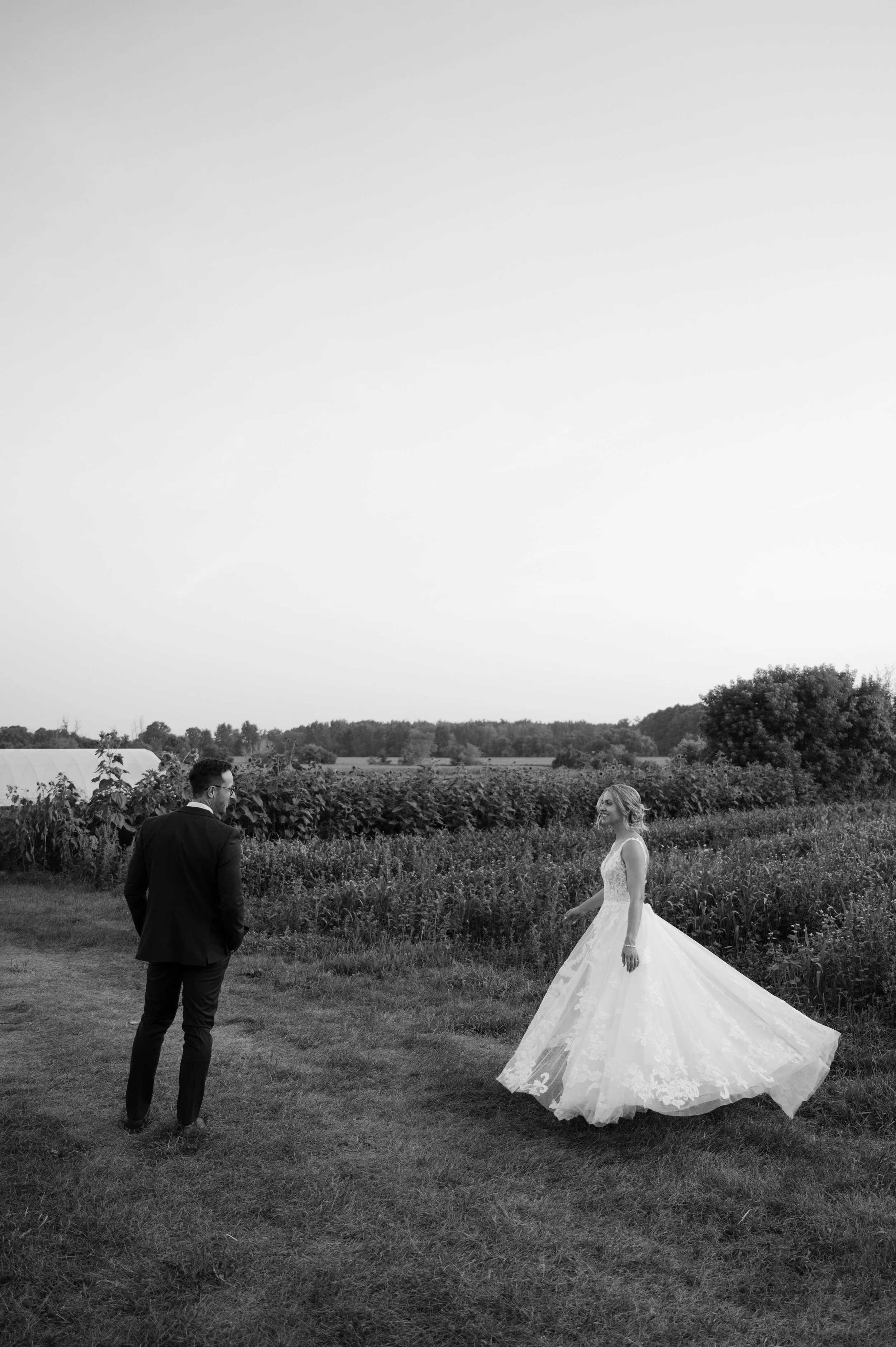 A bride in a wedding dress standing outside on grass, smiling at a man in a suit who is facing her. They are in an open field with trees and farmland in the background, under a clear sky.