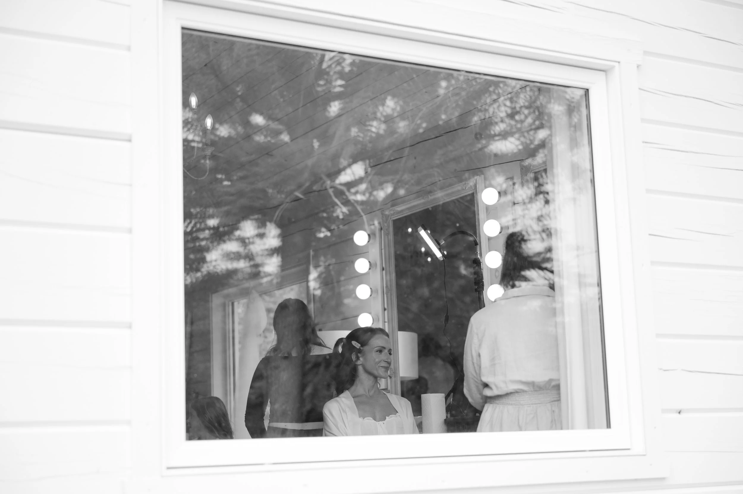 Black and white photo of a woman sitting in front of a mirror with light bulbs, getting her makeup done, with a person standing behind her, all reflected in the window outside.