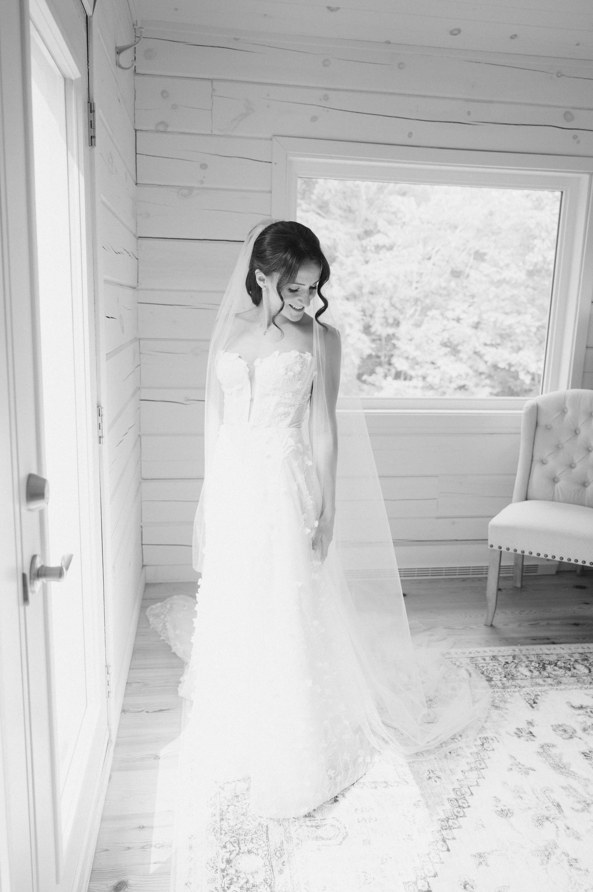 A bride in a wedding dress standing indoors near a window, smiling and looking down.