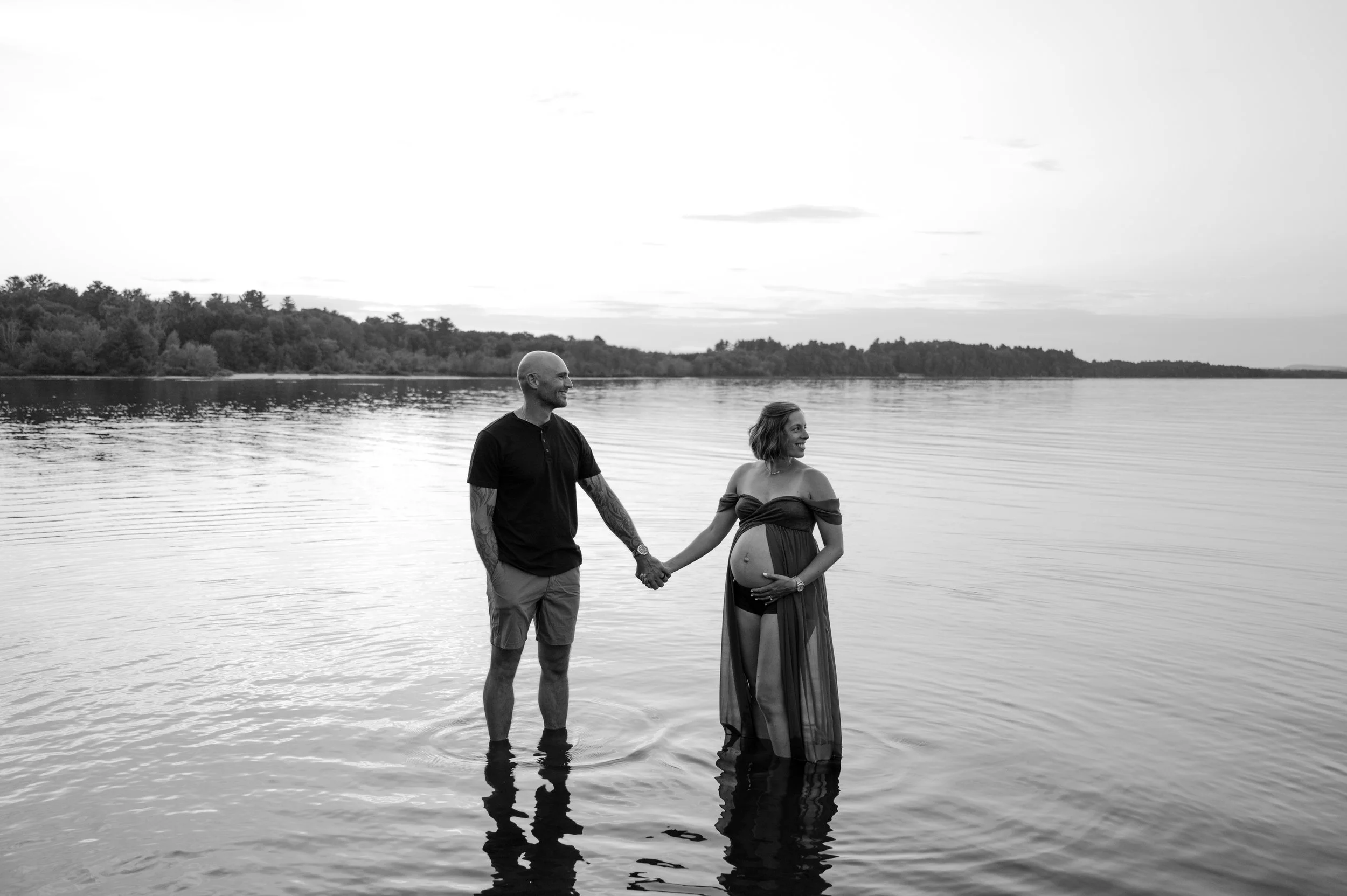 A couple standing in water holding hands during a photo shoot, the woman is pregnant and wearing a sheer cover-up, with a scenic lake and trees in the background.
