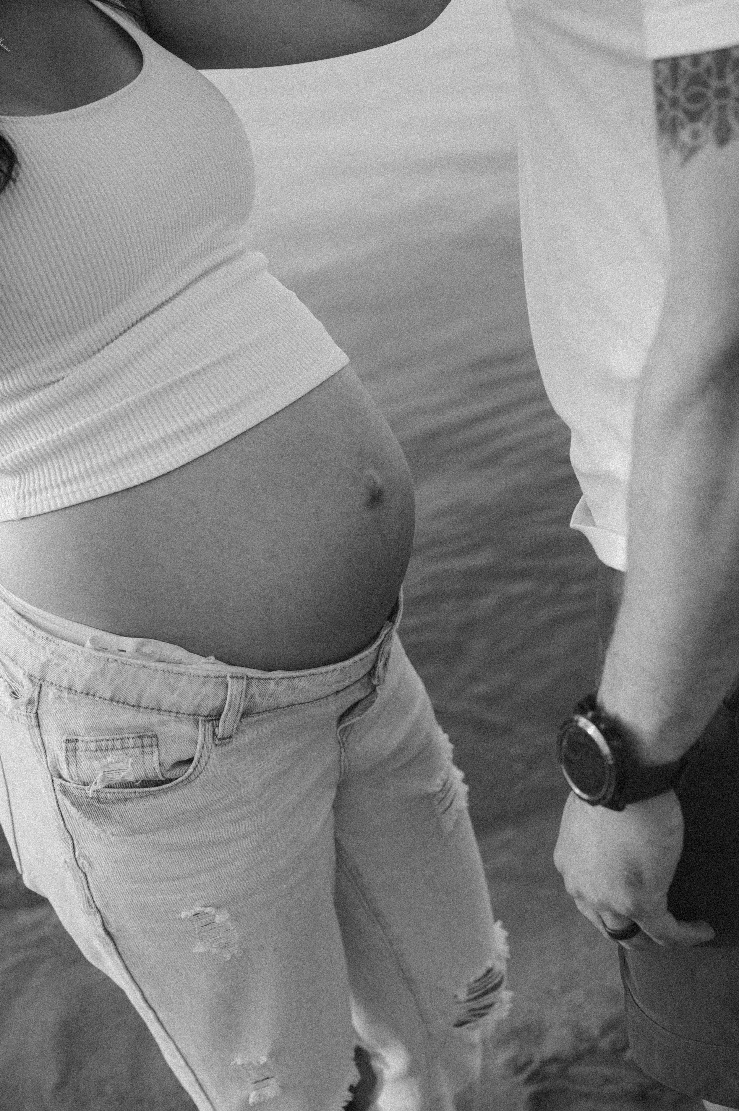 Close-up of a pregnant woman in casual clothing standing near water, with a man beside her wearing a watch.