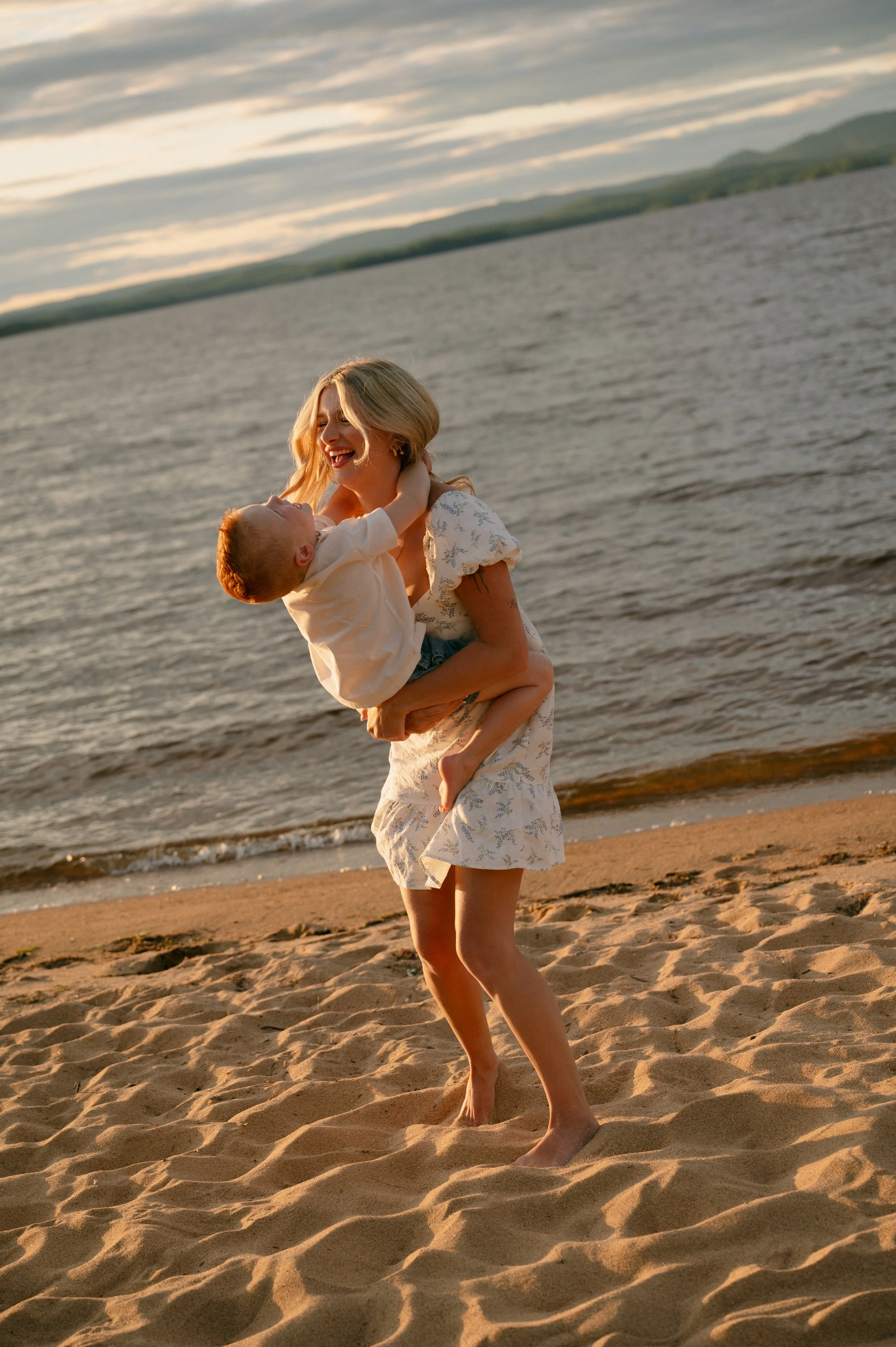 A woman playing with a young child on a sandy beach during sunset, with the ocean and distant hills in the background.