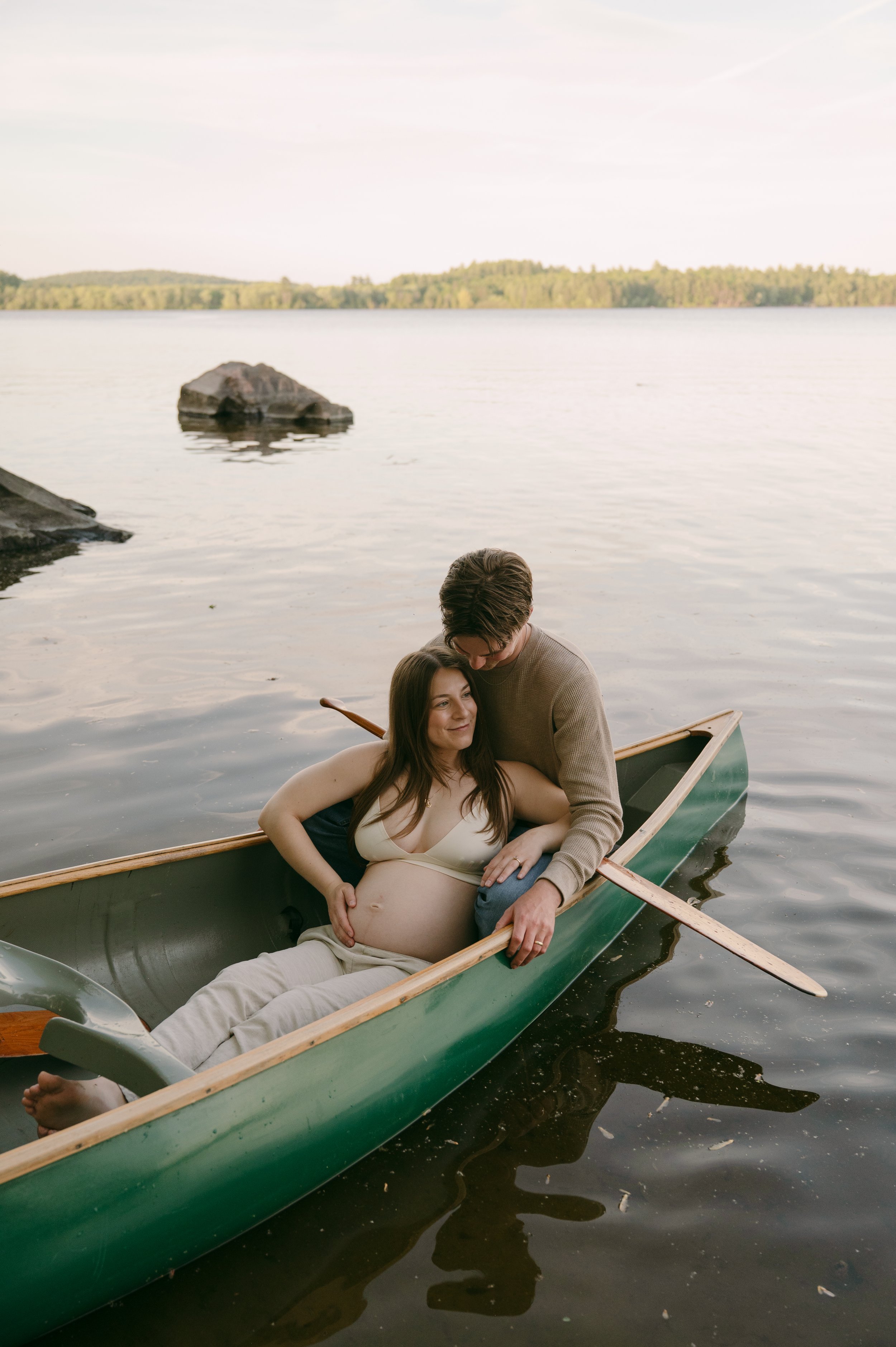 A pregnant woman lying in a canoe on water, with a man leaning over her, holding her belly, in a peaceful lakeside setting.