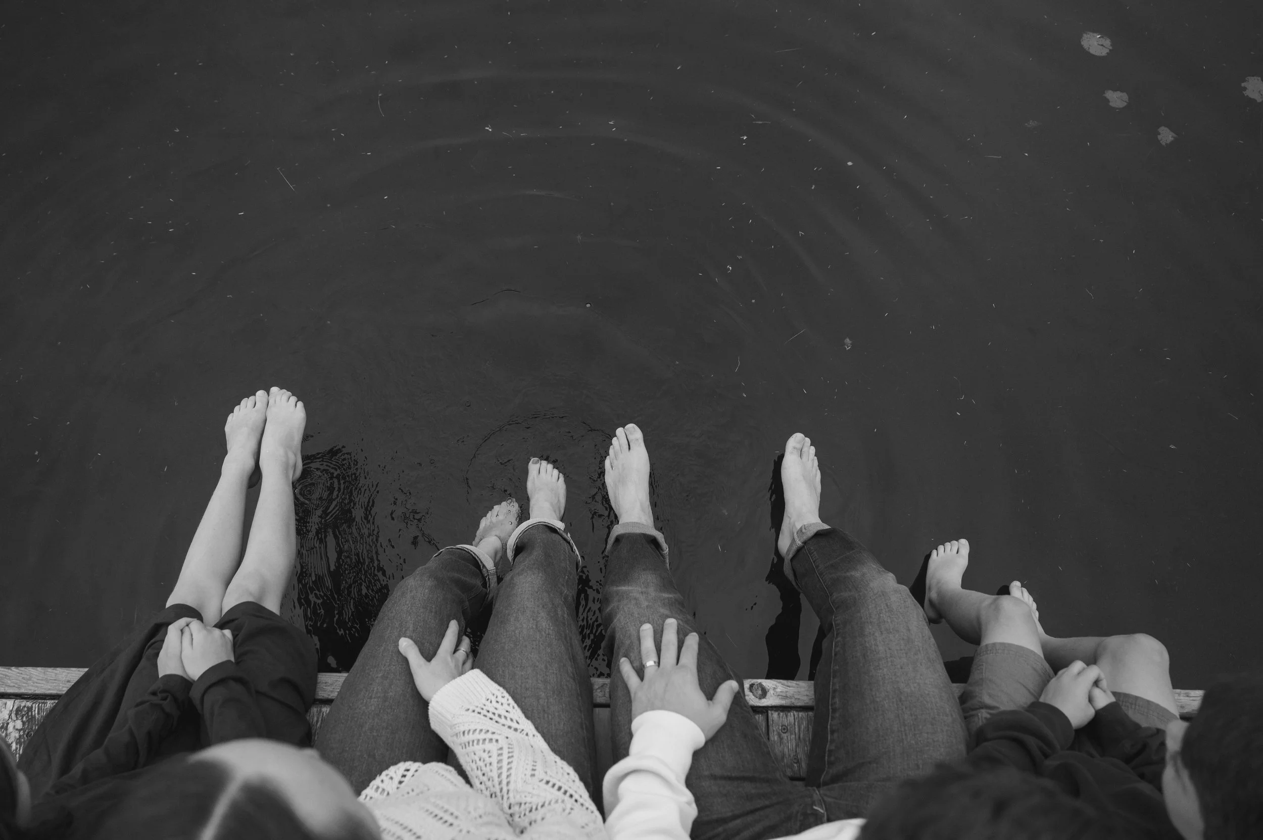 Four people with bare feet sitting on a dock with their feet in the water.