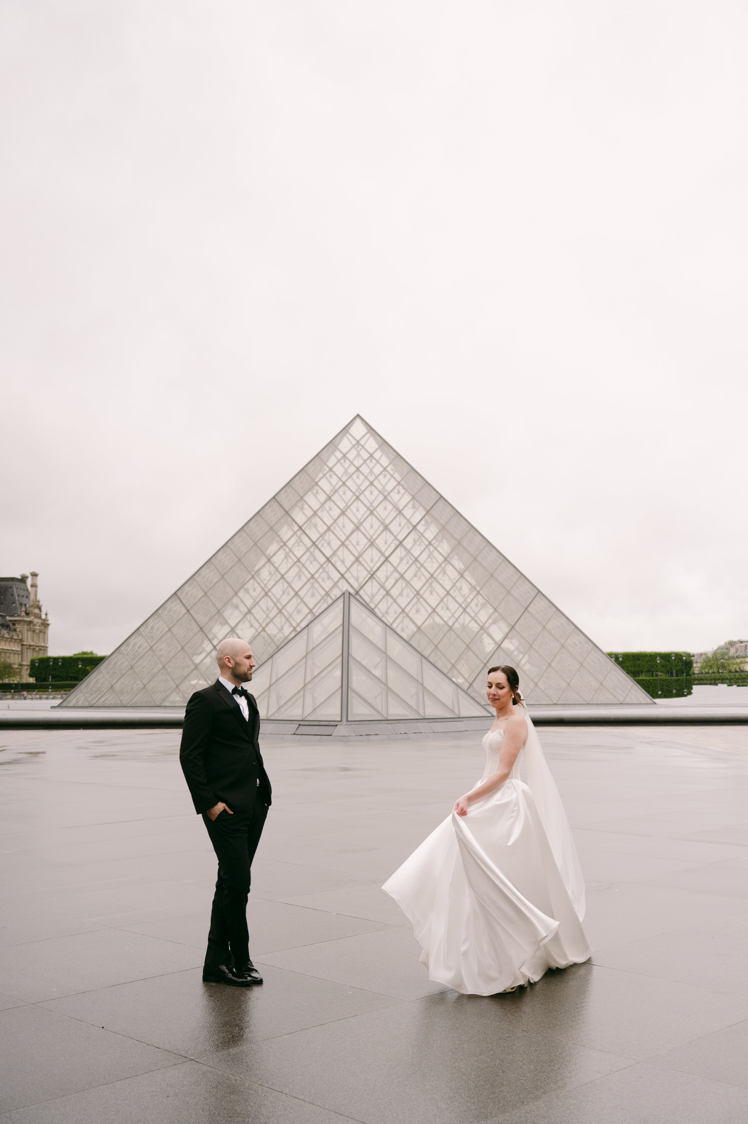 A bride and groom in wedding attire standing in front of the Louvre Pyramid in Paris on a cloudy day.