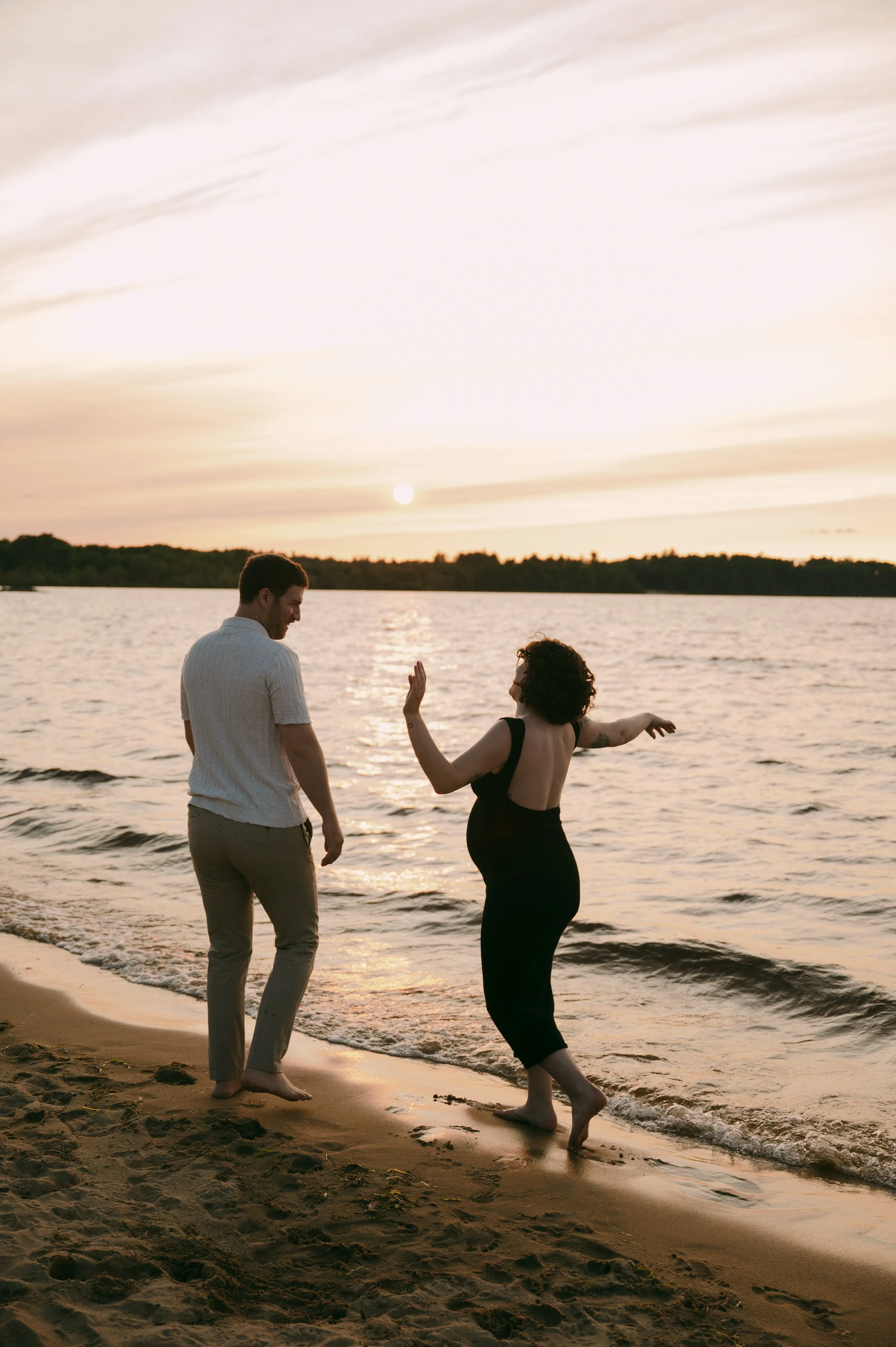 A man and pregnant woman dancing on the beach at sunset, with the ocean and a distant treeline in the background.