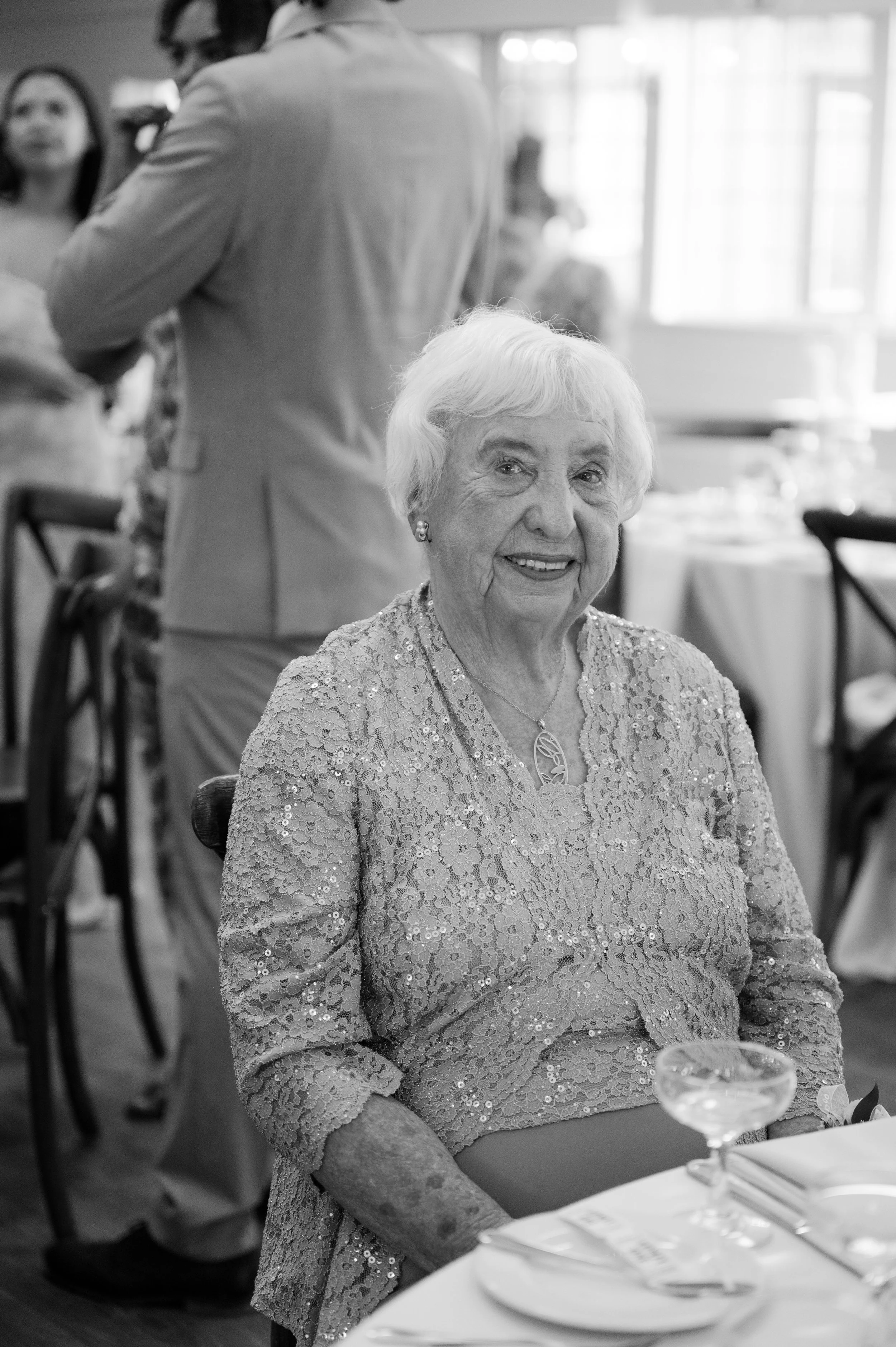 A smiling elderly woman with short white hair, wearing a sequined lace dress, seated at a table with place settings, in a festive restaurant or banquet setting.