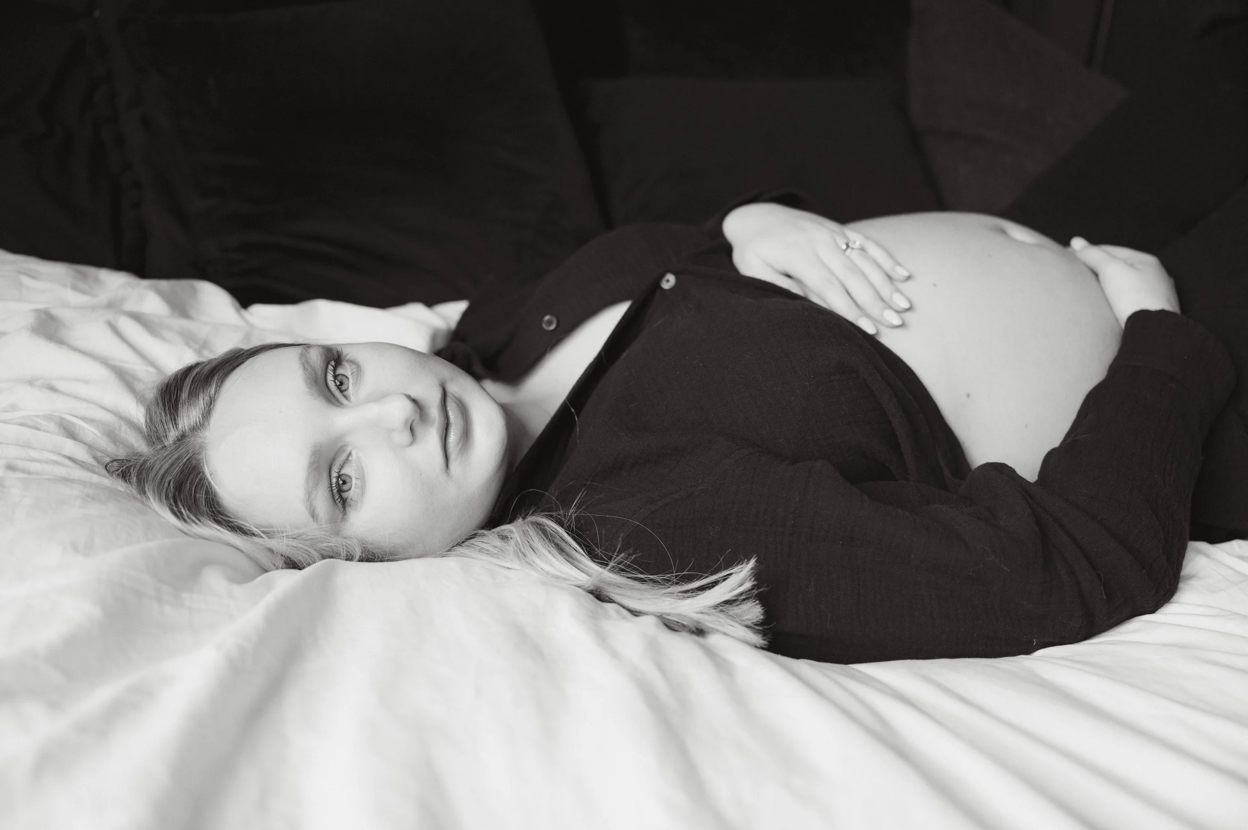 A pregnant woman lying on a bed with her hand resting on her stomach, looking at the camera.