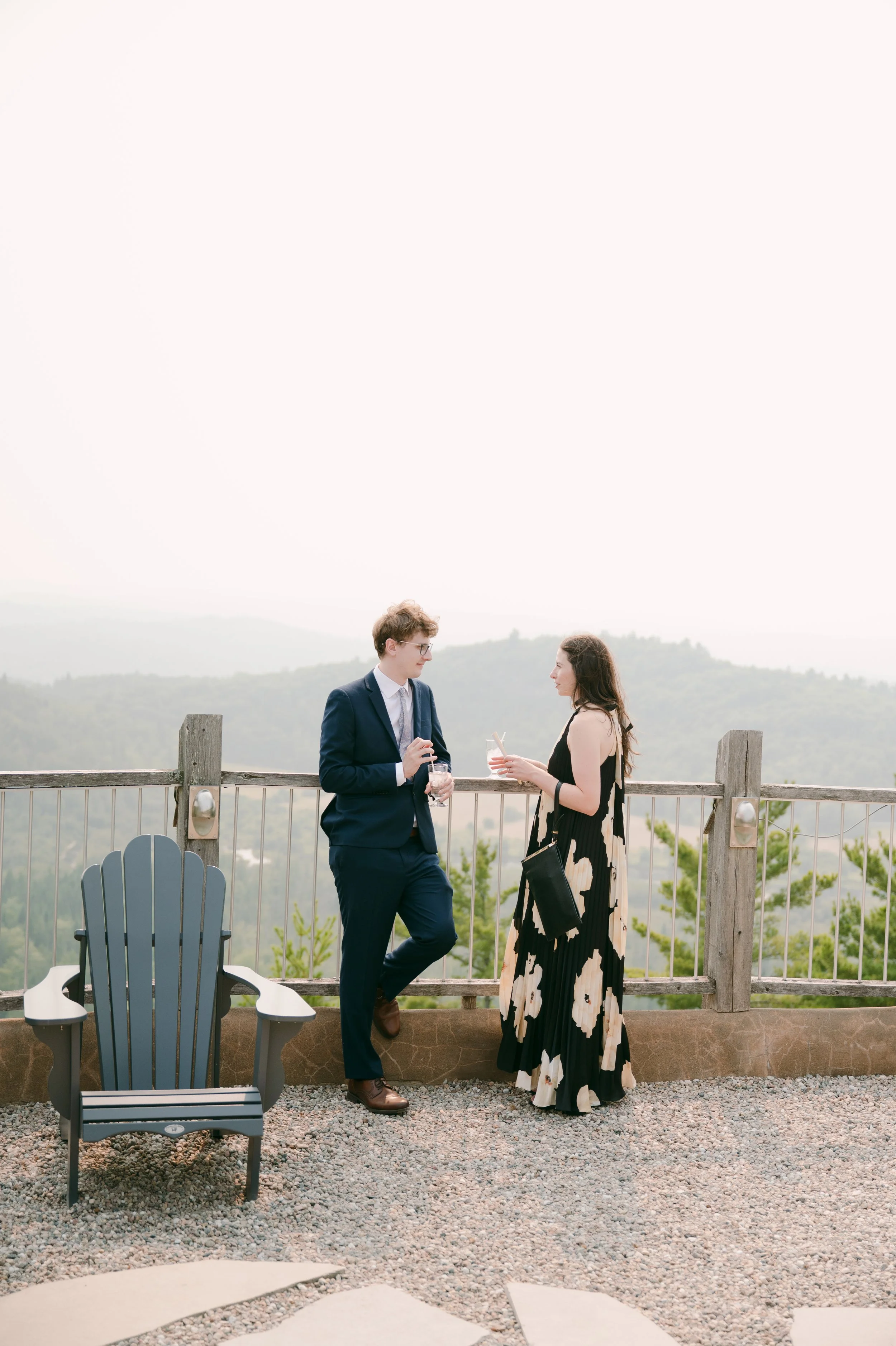 A man in a navy suit and a woman in a black floral dress are standing and talking on a balcony overlooking a hilly landscape. They are holding drinks.