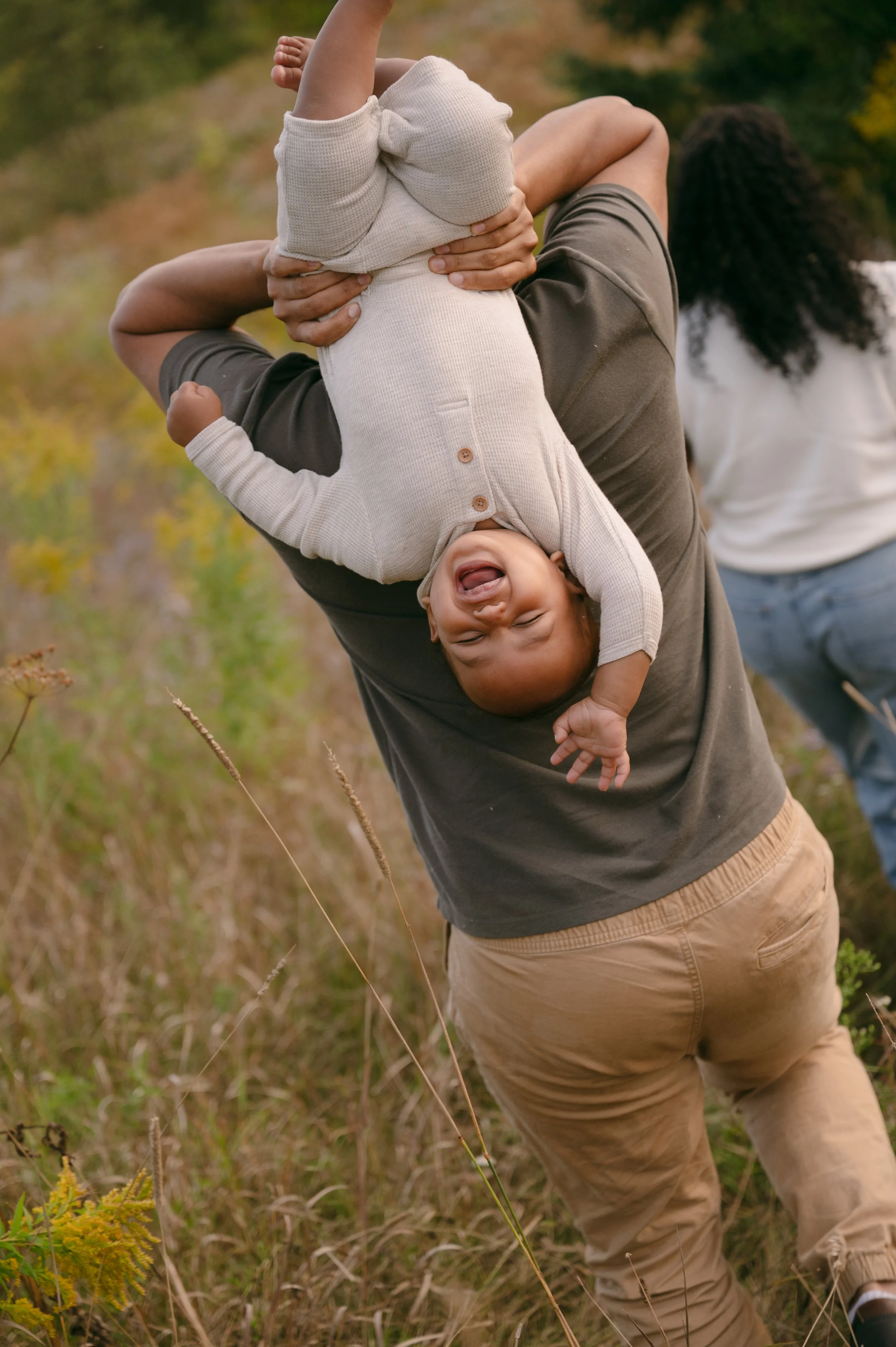 A man holding a young child upside down outdoors in a grassy field; the child is smiling and laughing, wearing a light-colored sweater and pants, with a woman in a white shirt and jeans standing in the background.