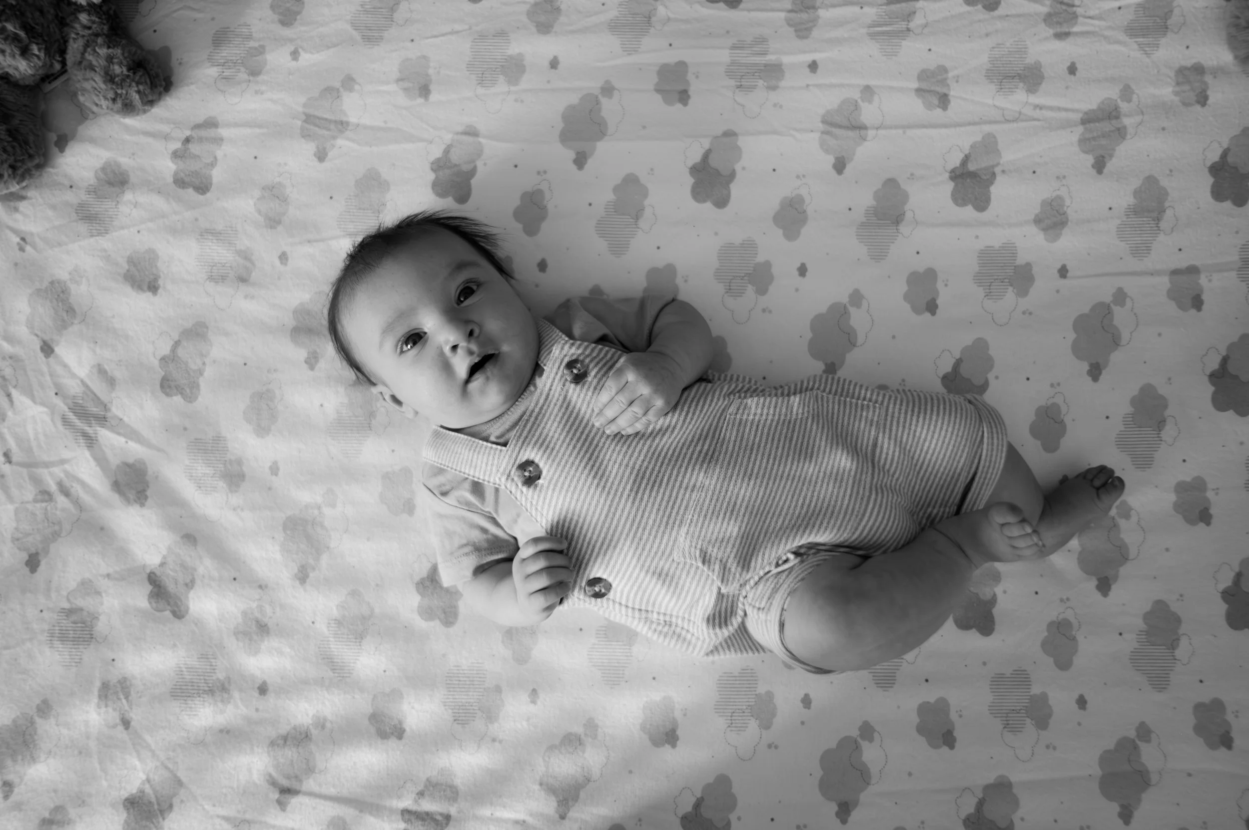 A baby lying on a sheet with a floral pattern, wearing a striped outfit, looking up at the camera with a curious expression.