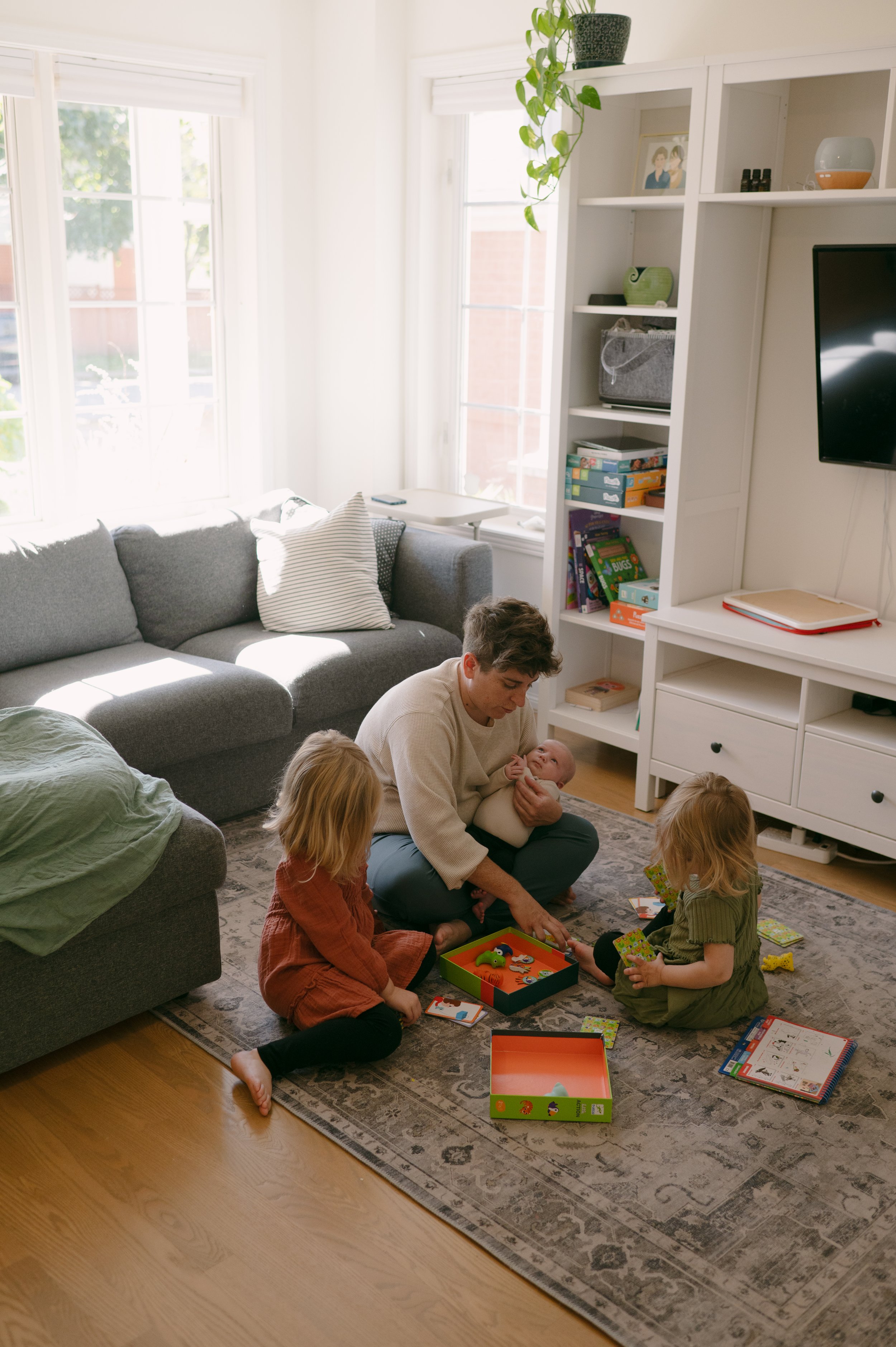 A family sitting on a patterned rug in a living room, playing a board game with three children and an adult holding an infant, near a gray sofa and white bookshelf with various books and decor.