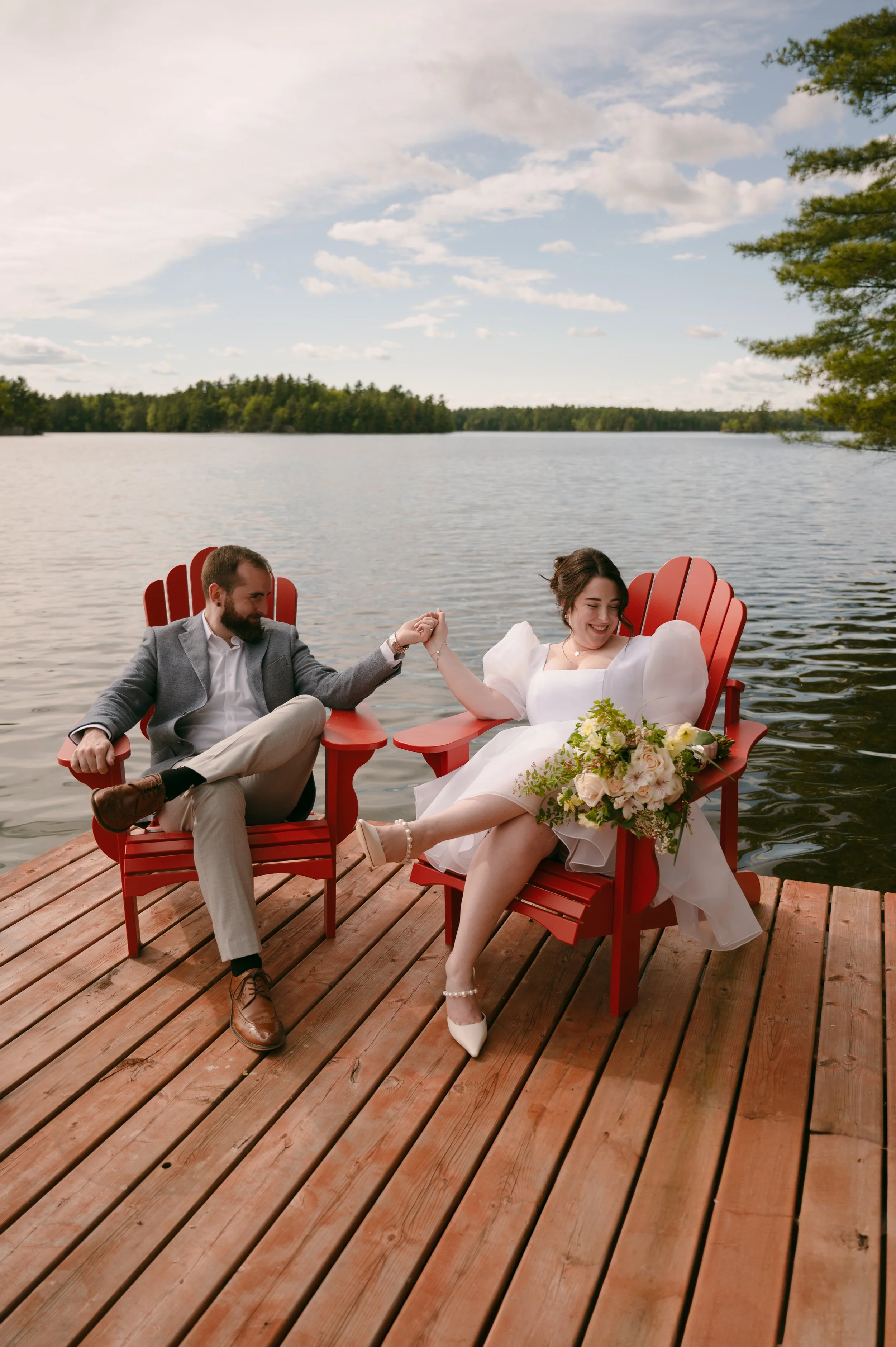 A bride and groom sitting on a wooden dock by a lake, holding hands, with the bride smiling, holding a bouquet, and the groom looking at her, both seated on red Adirondack chairs.