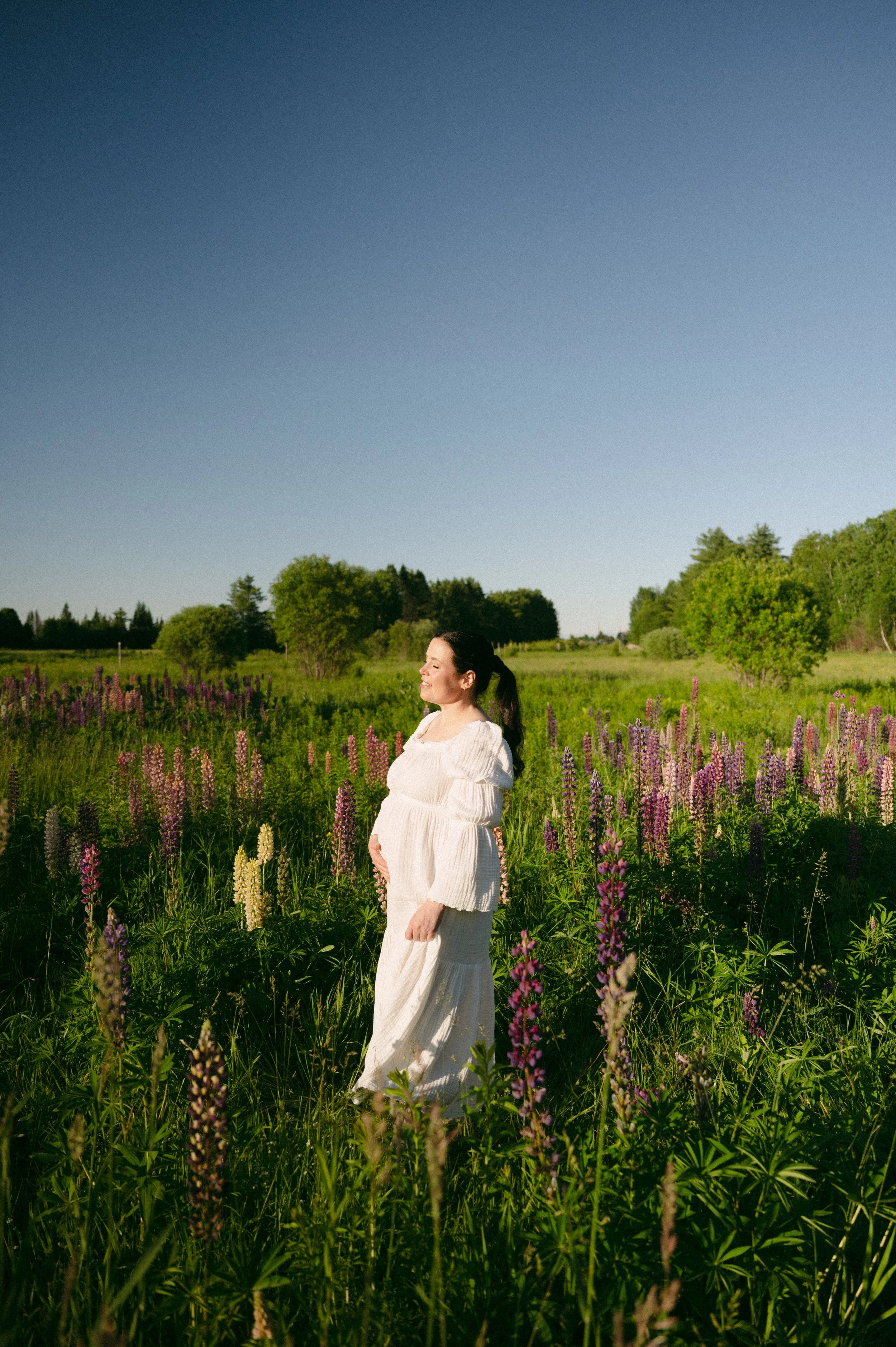 A woman in a white dress standing in a field of purple and yellow flowers, smiling with eyes closed, under a clear blue sky.