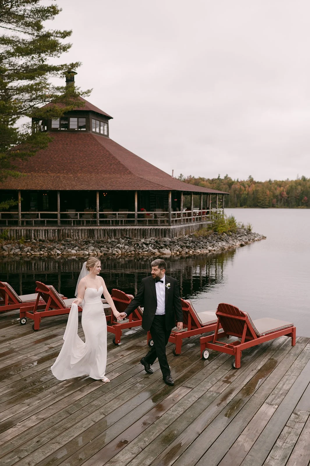 A newlywed couple walking hand in hand on a wooden dock by a lake. The bride is wearing a white wedding gown, and the groom is dressed in a black tuxedo. Old red lounge chairs are arranged on the dock, and a large, round wooden building with a red roof is in the background.