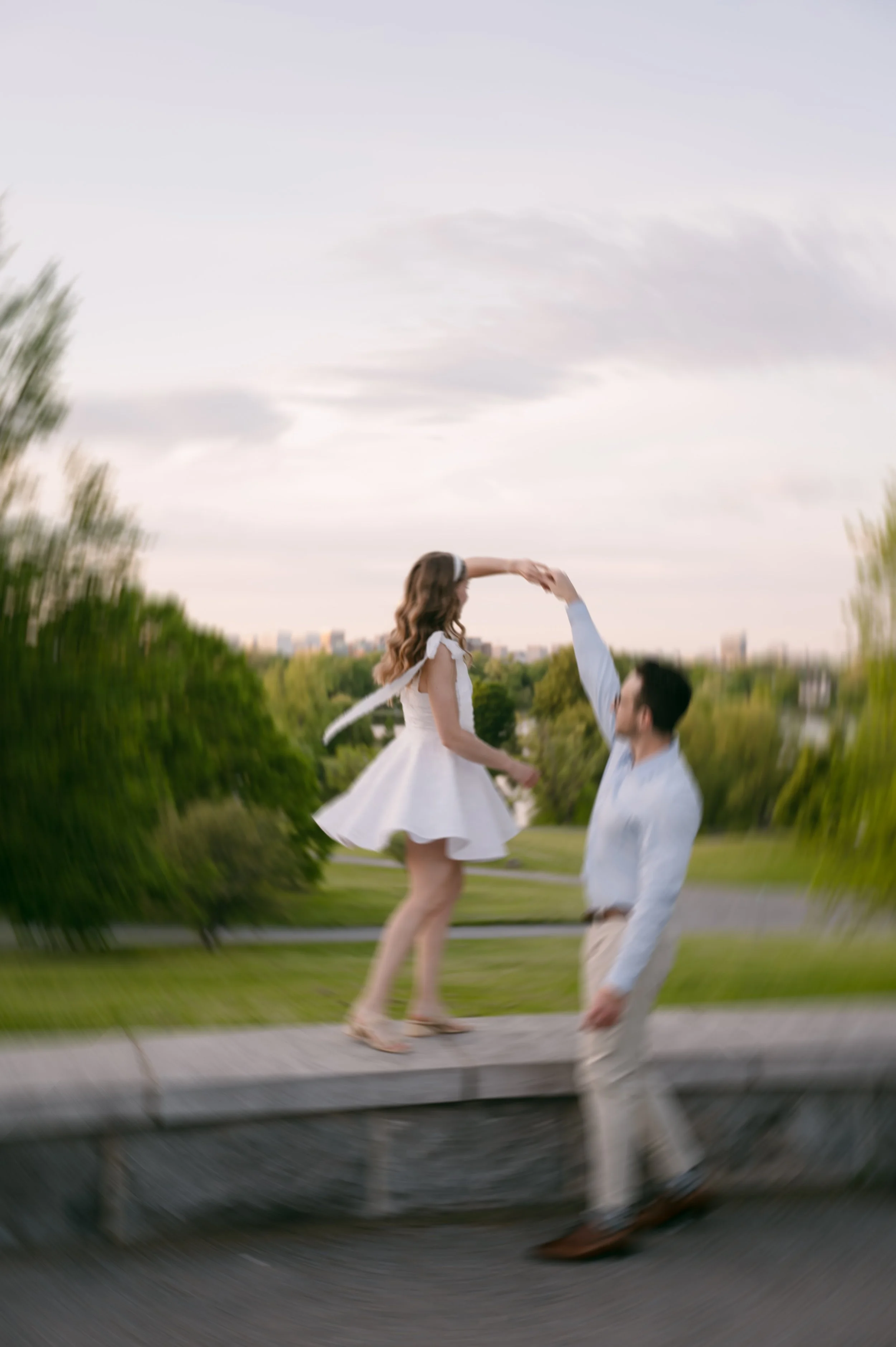 A couple dancing outdoors on a park pathway at sunset, with the woman standing on a raised surface and the man holding her hand. The woman wears a white dress, and the man wears a white shirt and beige pants.