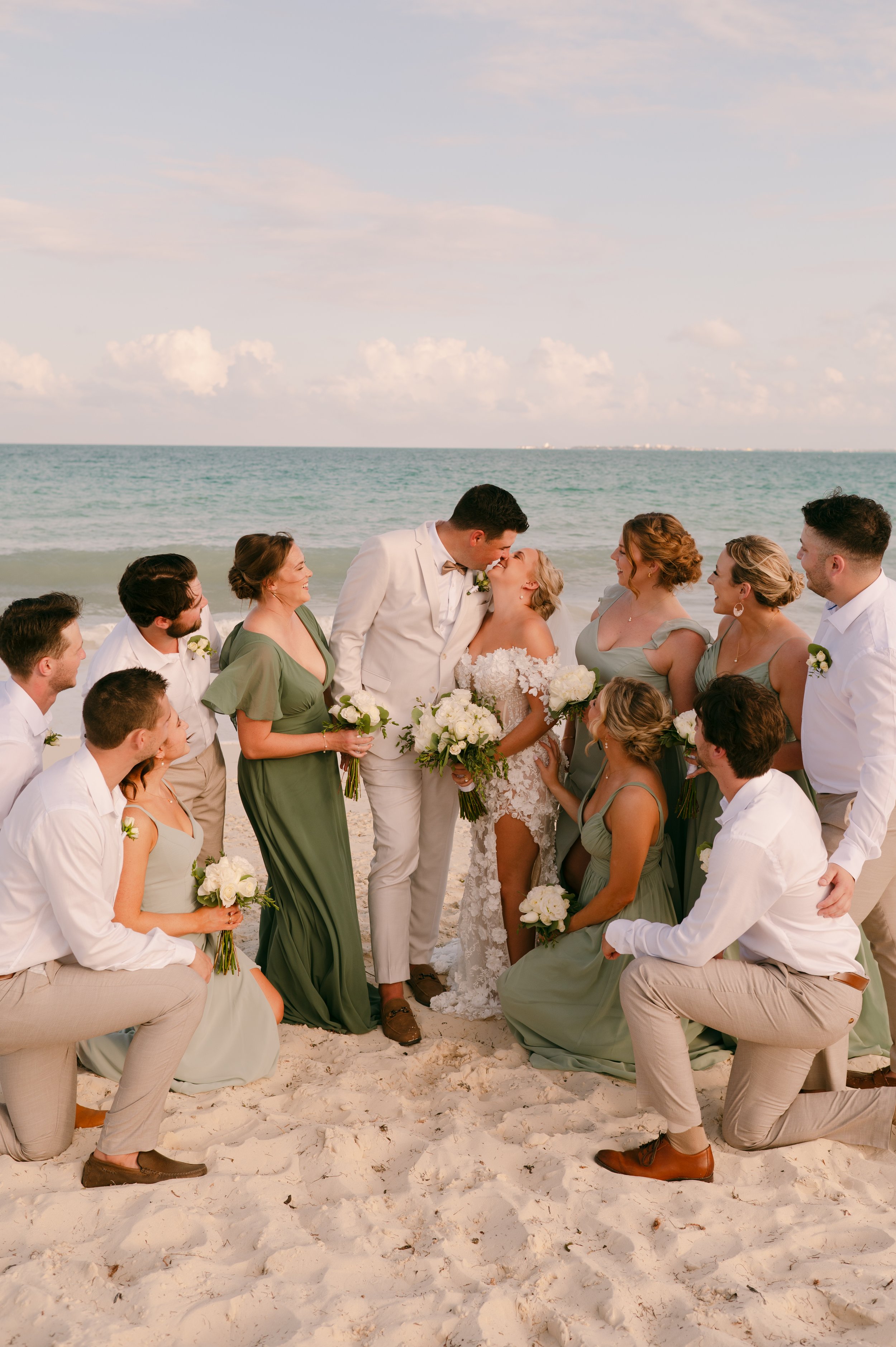 A wedding party on the beach, with a groom and bride kissing in the center, surrounded by their wedding guests holding bouquets, with the ocean and sky in the background.