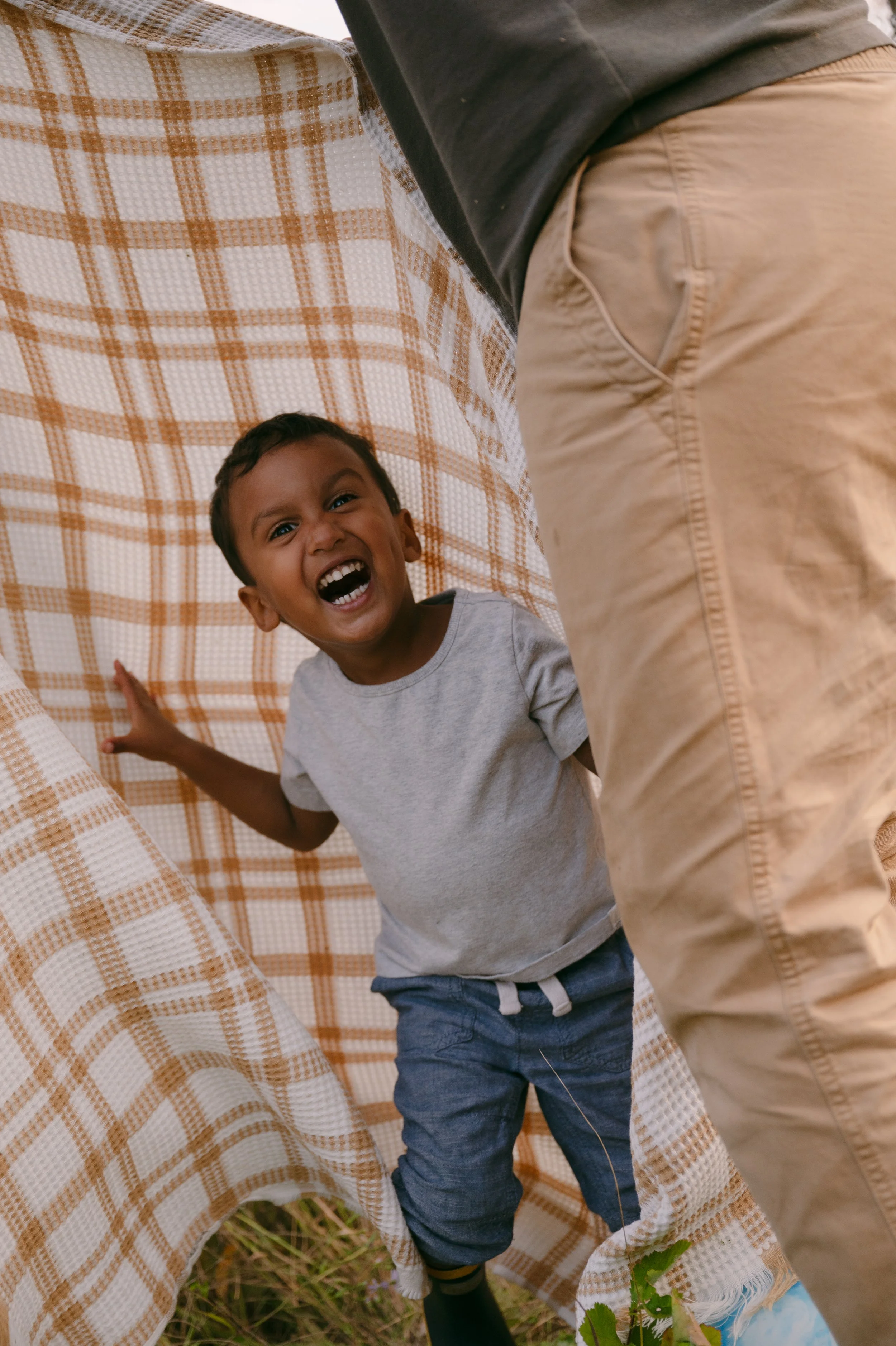 A young boy with dark hair and a big smile, wearing a gray t-shirt and blue shorts, standing behind a checkered cloth tent outdoors, with part of an adult's leg visible in the foreground.
