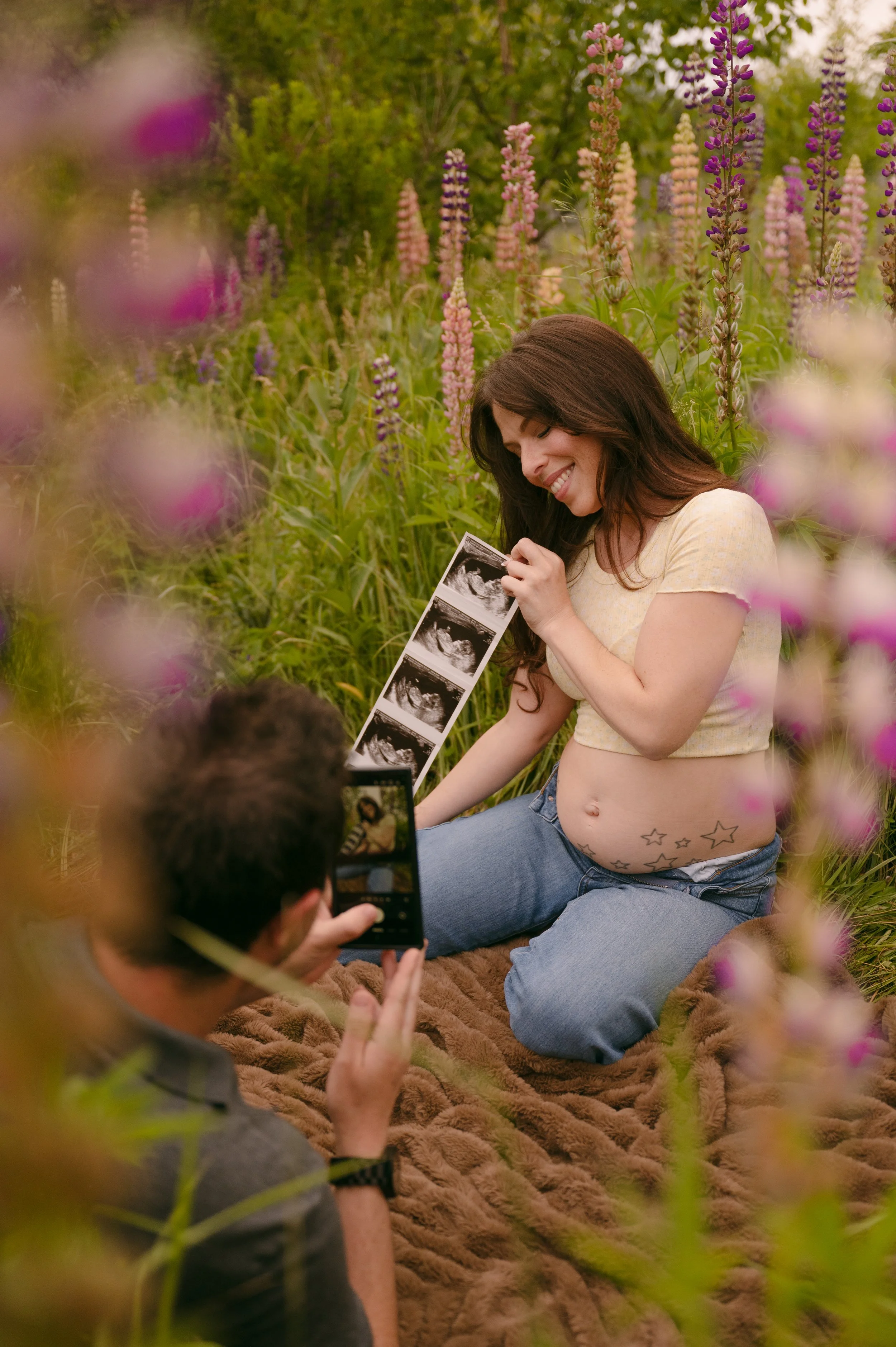 Pregnant woman sitting on a blanket in a field of tall pink and purple flowers, smiling as she looks at ultrasound images, with a person taking her photo.