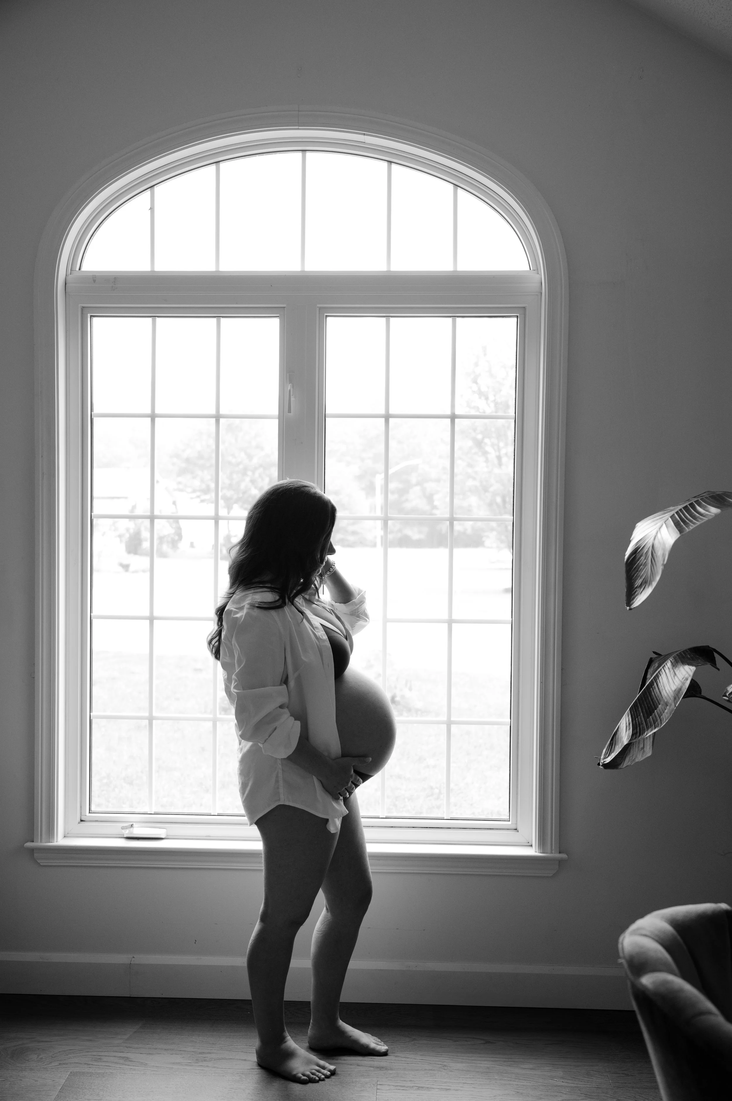 A pregnant woman standing barefoot inside a room with a large window, looking out while holding her belly.