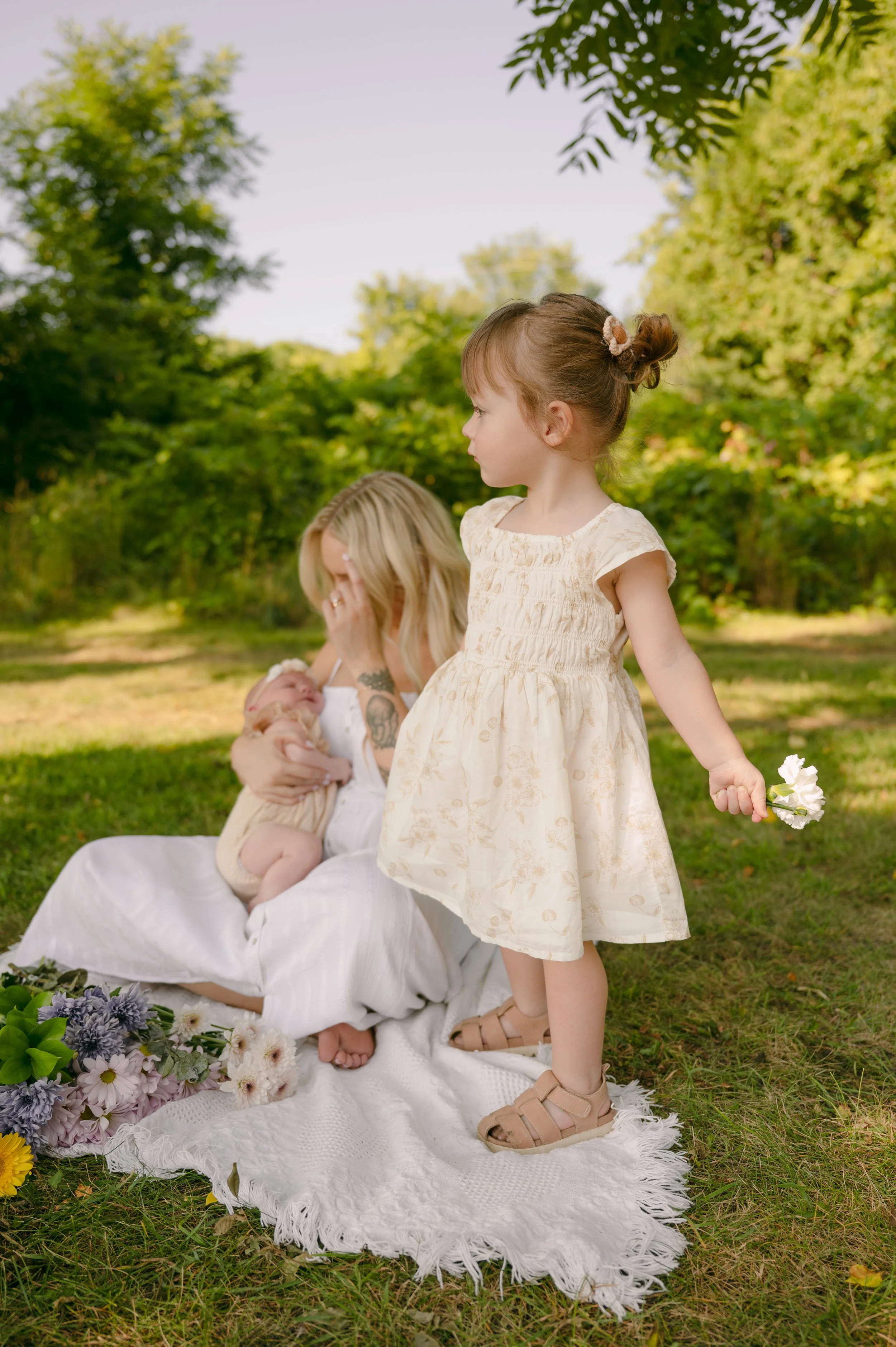 A young girl holding a flower stands next to a woman and a baby, sitting on a white blanket outdoors in a park with lush green trees in the background.