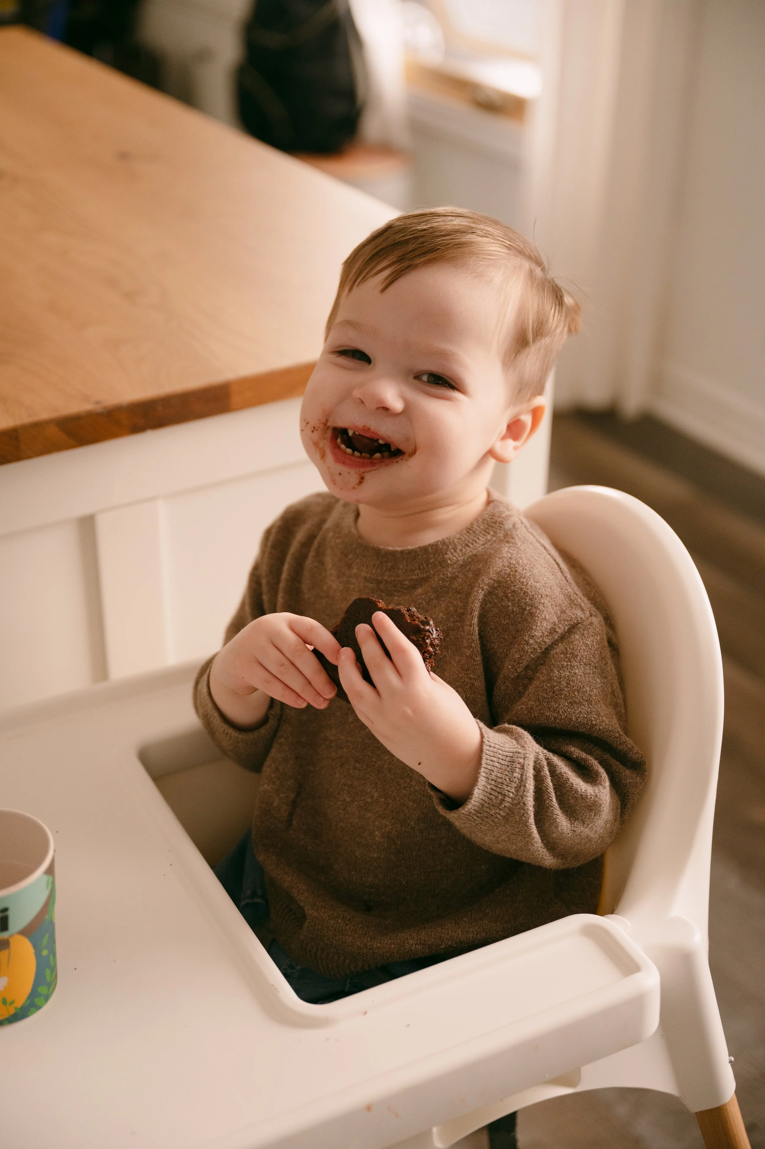 A young child sitting in a high chair, holding a piece of chocolate cake, smiling with chocolate on his face, in a kitchen setting.