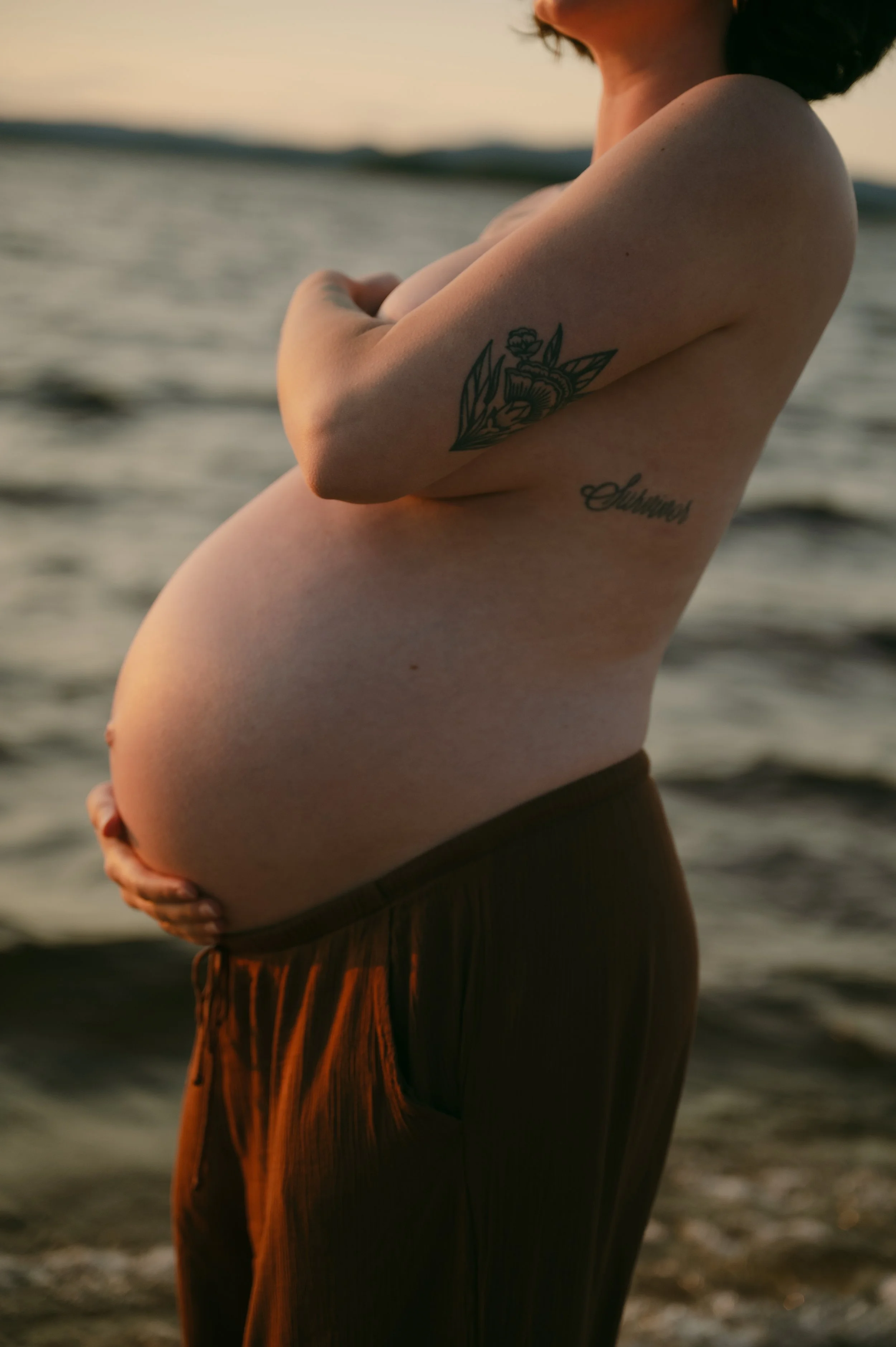 A pregnant woman standing by the water at sunset, holding her belly, with tattoos on her arm and side.