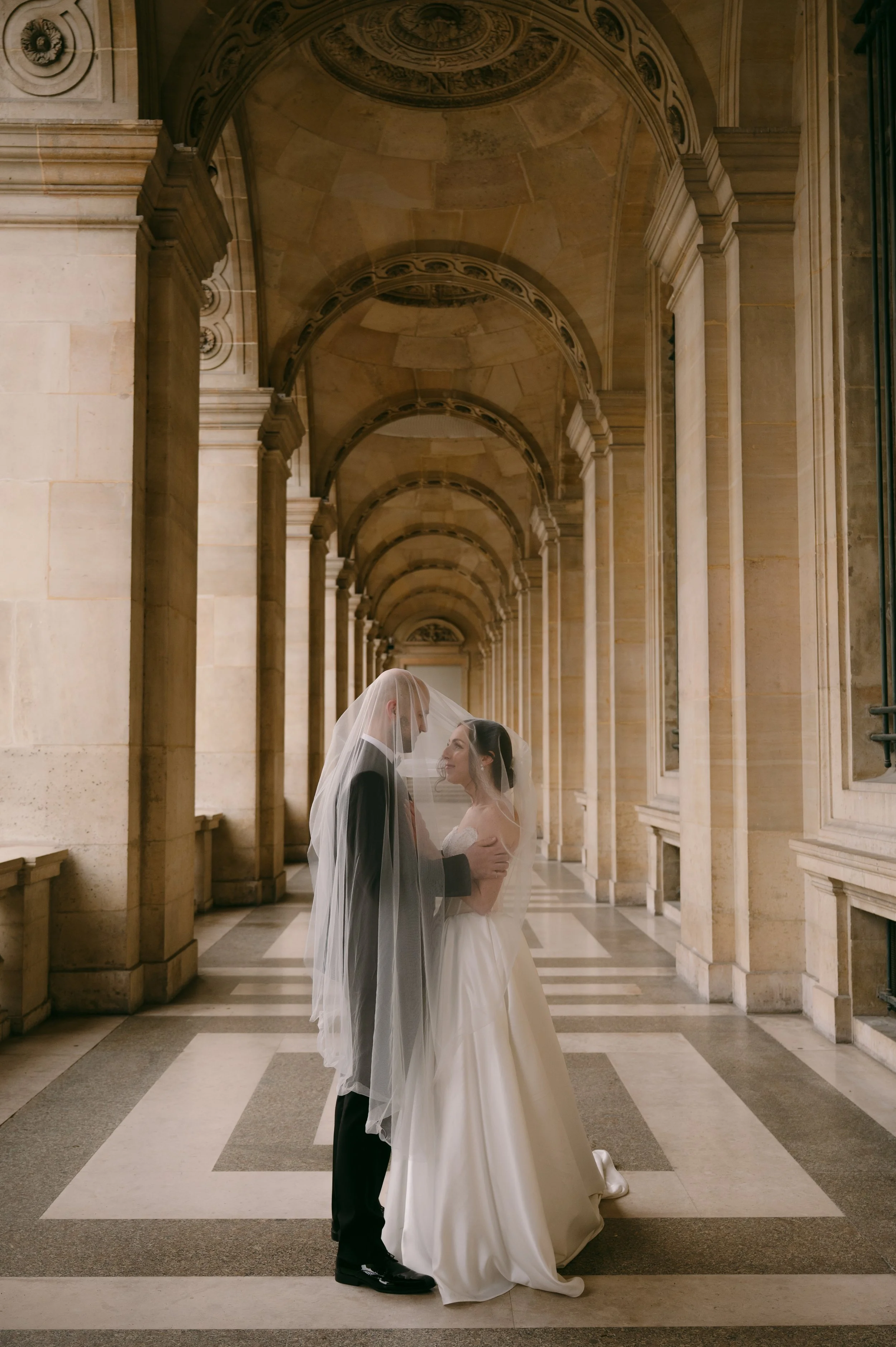 A bride and groom stand close together under a sheer veil in an elegant, arched hallway with stone columns and detailed ceiling murals.
