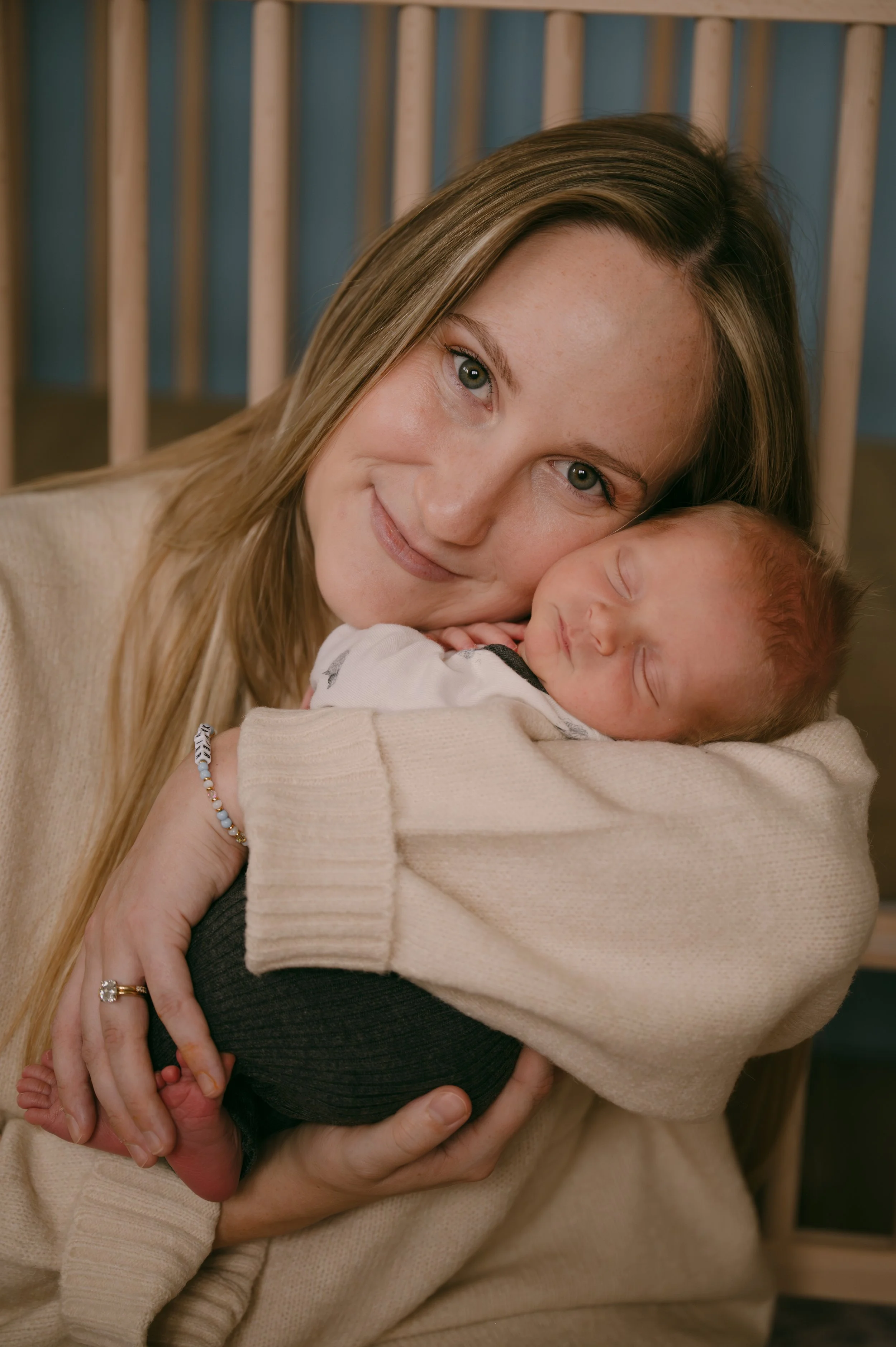 A woman with long blonde hair holding a sleeping newborn baby with freckles and light brown hair, smiling and resting her face on the baby's head.