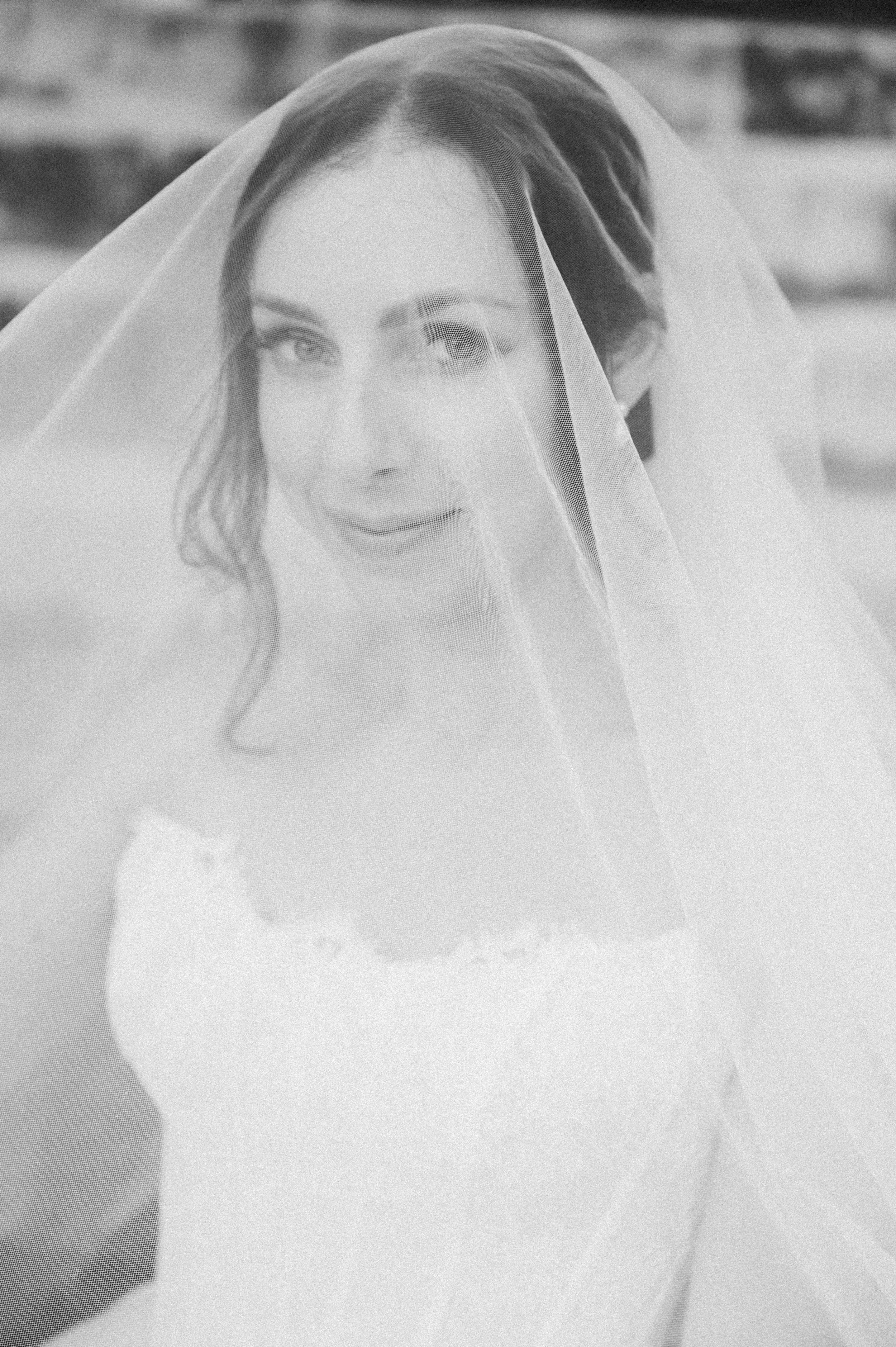A black-and-white photograph of a woman wearing a wedding veil and bra, with a blurred background.