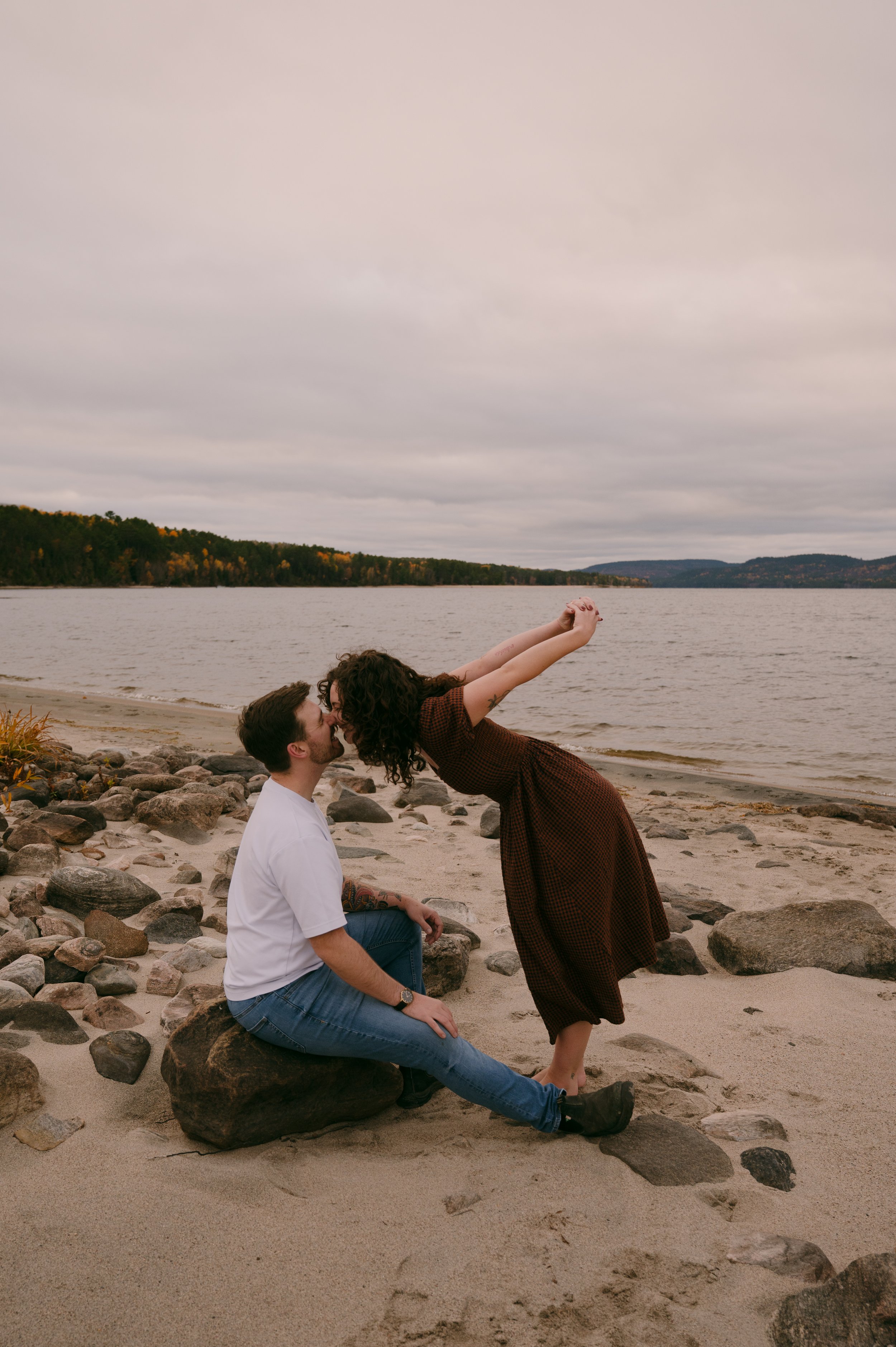A couple on a beach with rocks, where the woman is leaning over to kiss the man who is sitting on a large rock, with a lake and distant hills under a cloudy sky in the background.