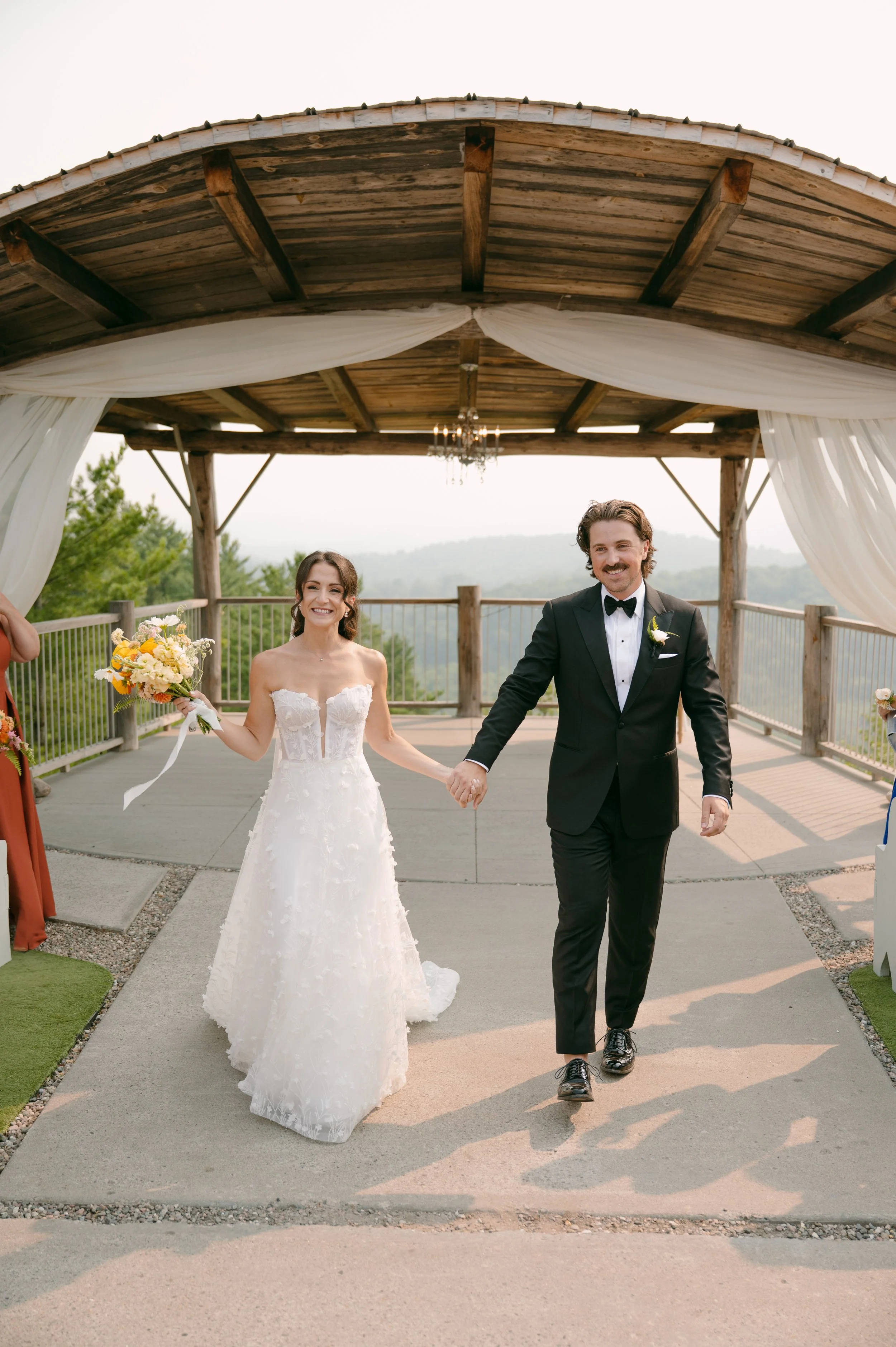 Happy bride and groom holding hands during their outdoor wedding ceremony, walking under a wooden pavilion with white drapes and a scenic mountain view in the background.