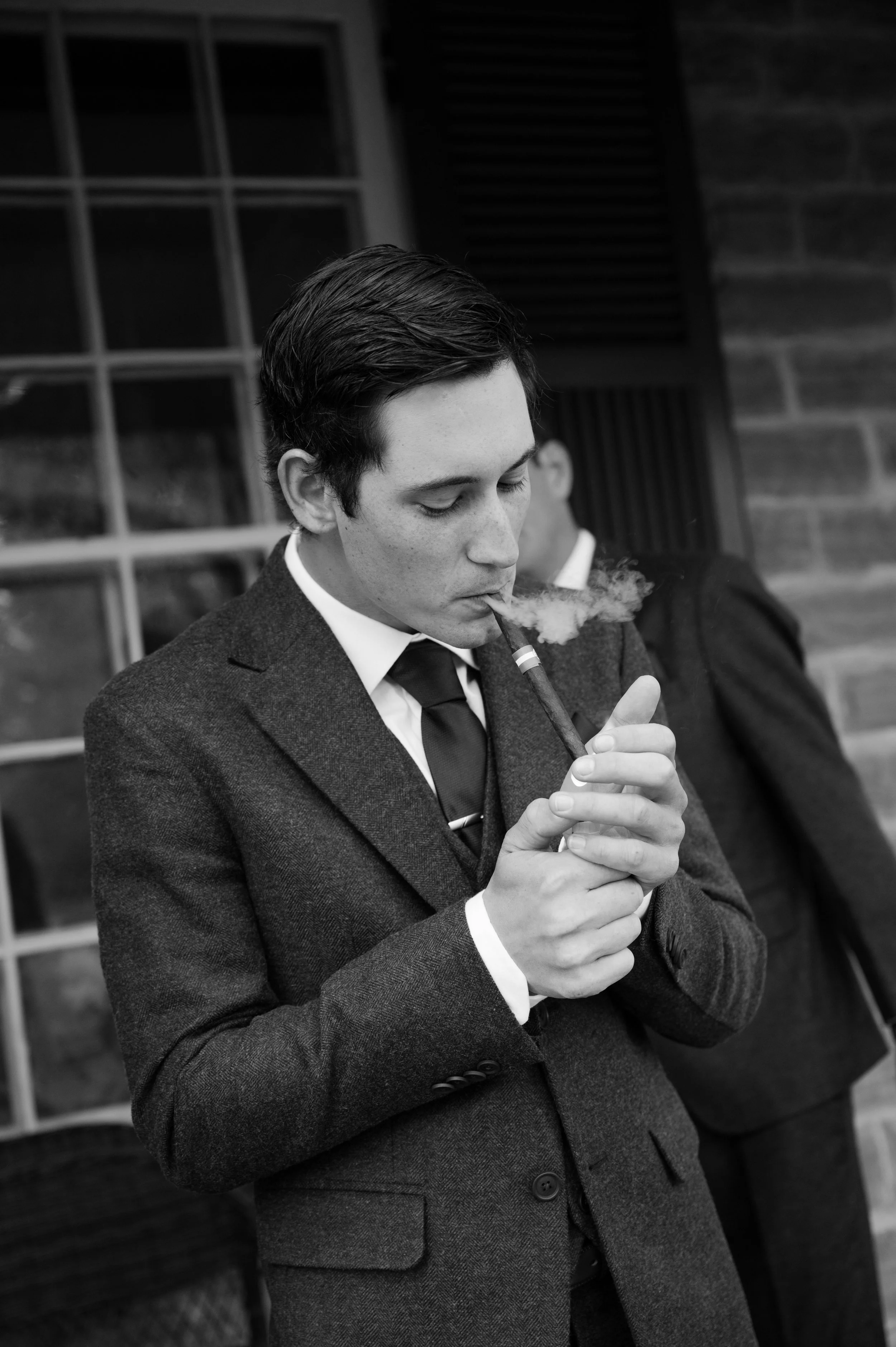 A young man with dark hair in a suit and tie, smoking a cigar outdoors near a brick building.