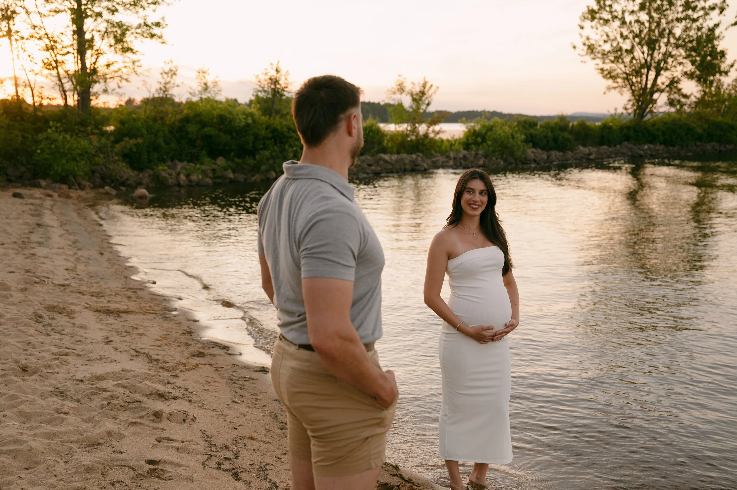 A pregnant woman in a white dress standing near a lake at sunset, smiling at a man in a gray shirt and beige shorts.