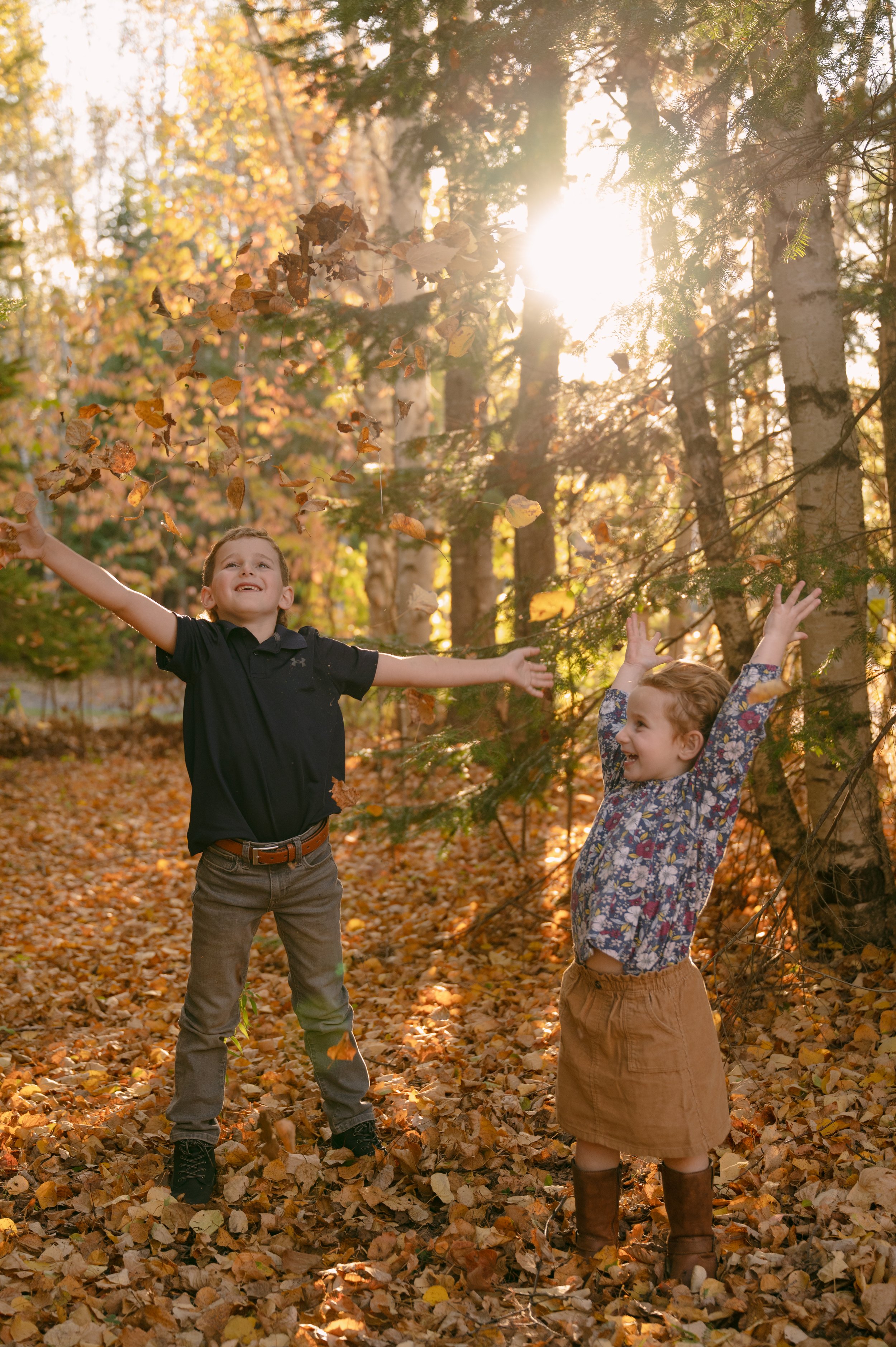 Two children playing in an autumn forest, throwing leaves in the air with sunlight filtering through trees.