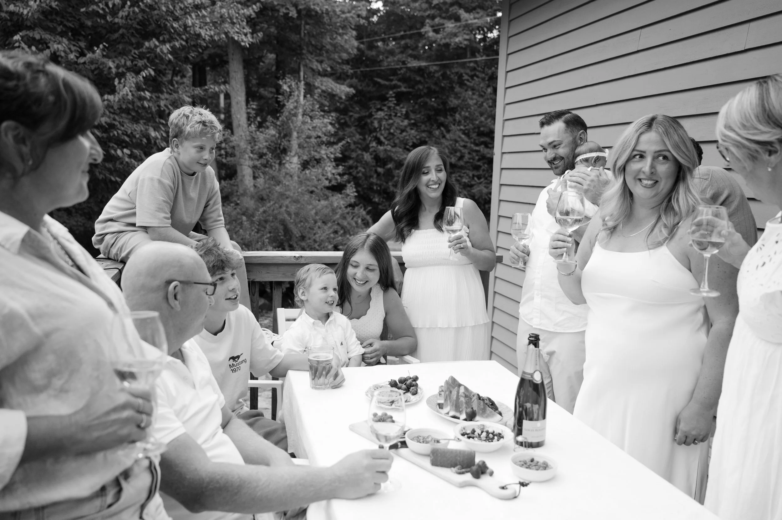 Family and friends gathered outdoors around a dining table, celebrating with drinks and food, with some standing and others sitting, in a wooded backyard setting.