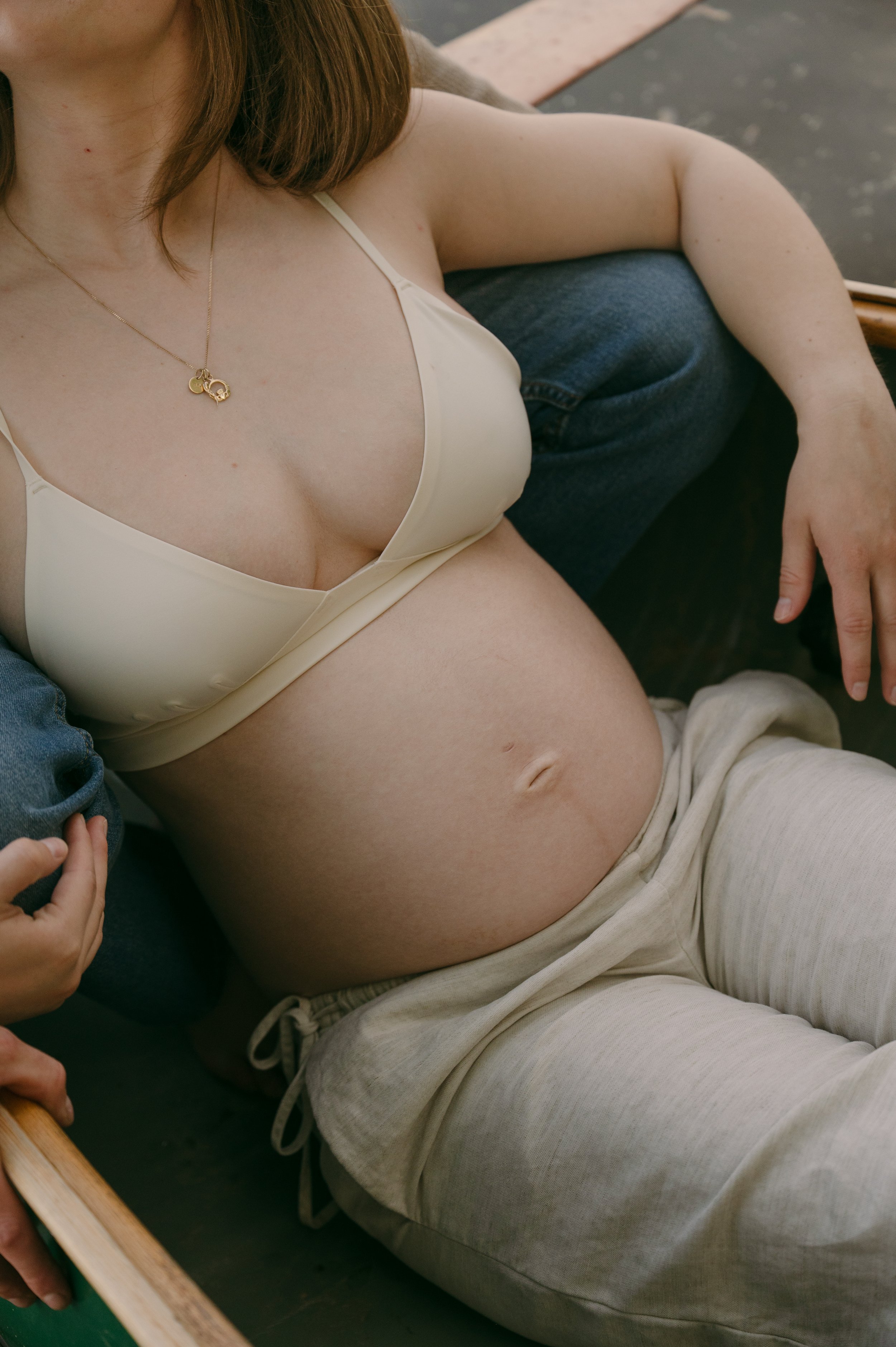 A person lying down on a wooden platform wearing a cream-colored bra, beige sweatpants, and a gold necklace with a heart-shaped pendant. The person's face is not visible, and they are holding a child's hand.